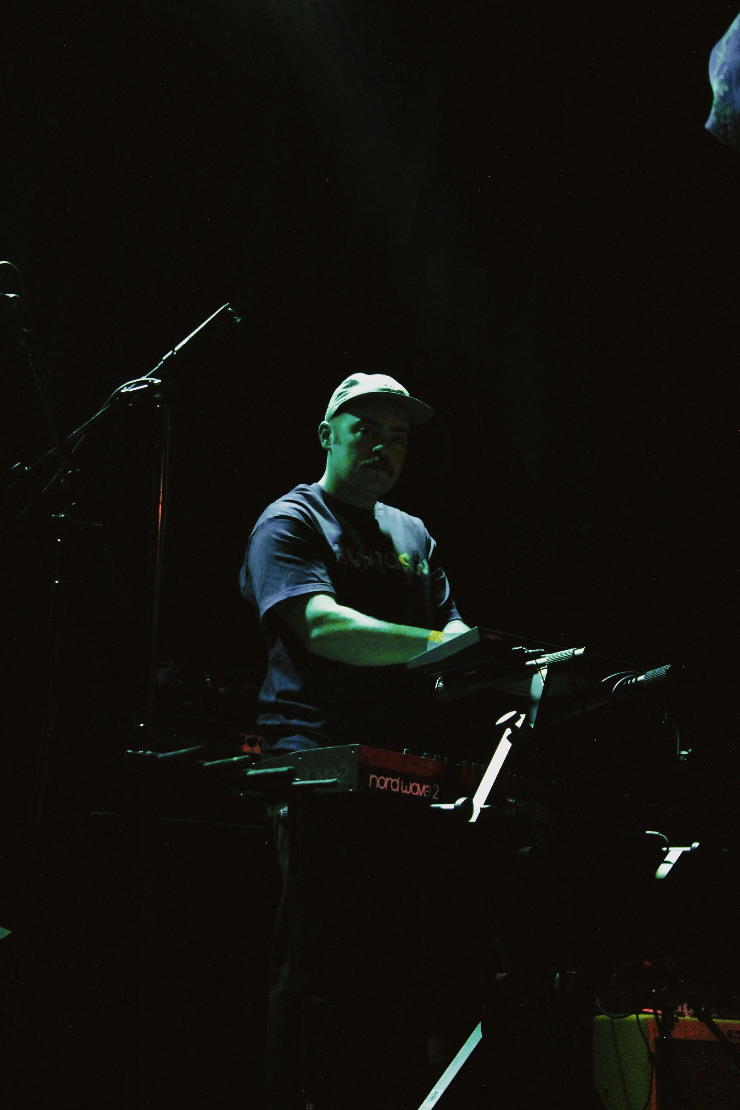 Musician playing keyboard on stage during a dark concert, wearing a cap and dark t-shirt.