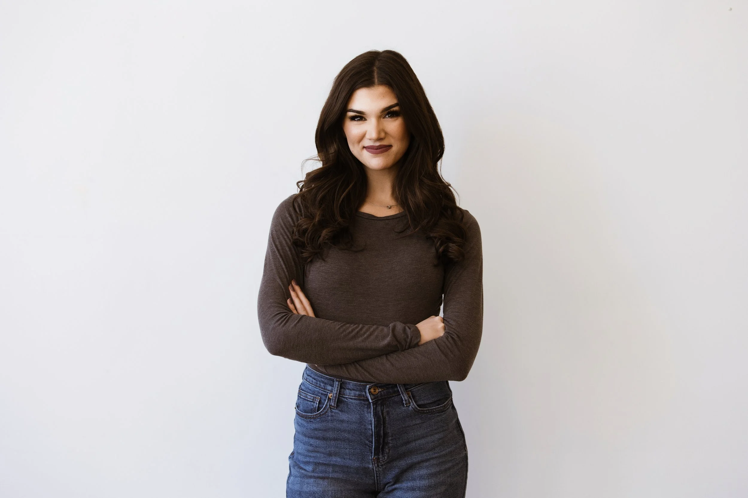 A young woman standing with her arms crossed, smiling at the camera, against a plain white wall.