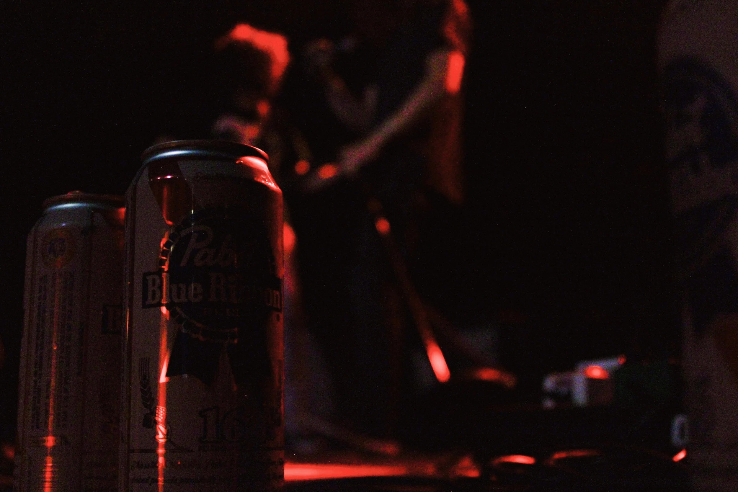 Two cans of Pabst Blue Ribbon beer in the foreground, with people talking in a dimly lit environment with red lighting in the background.