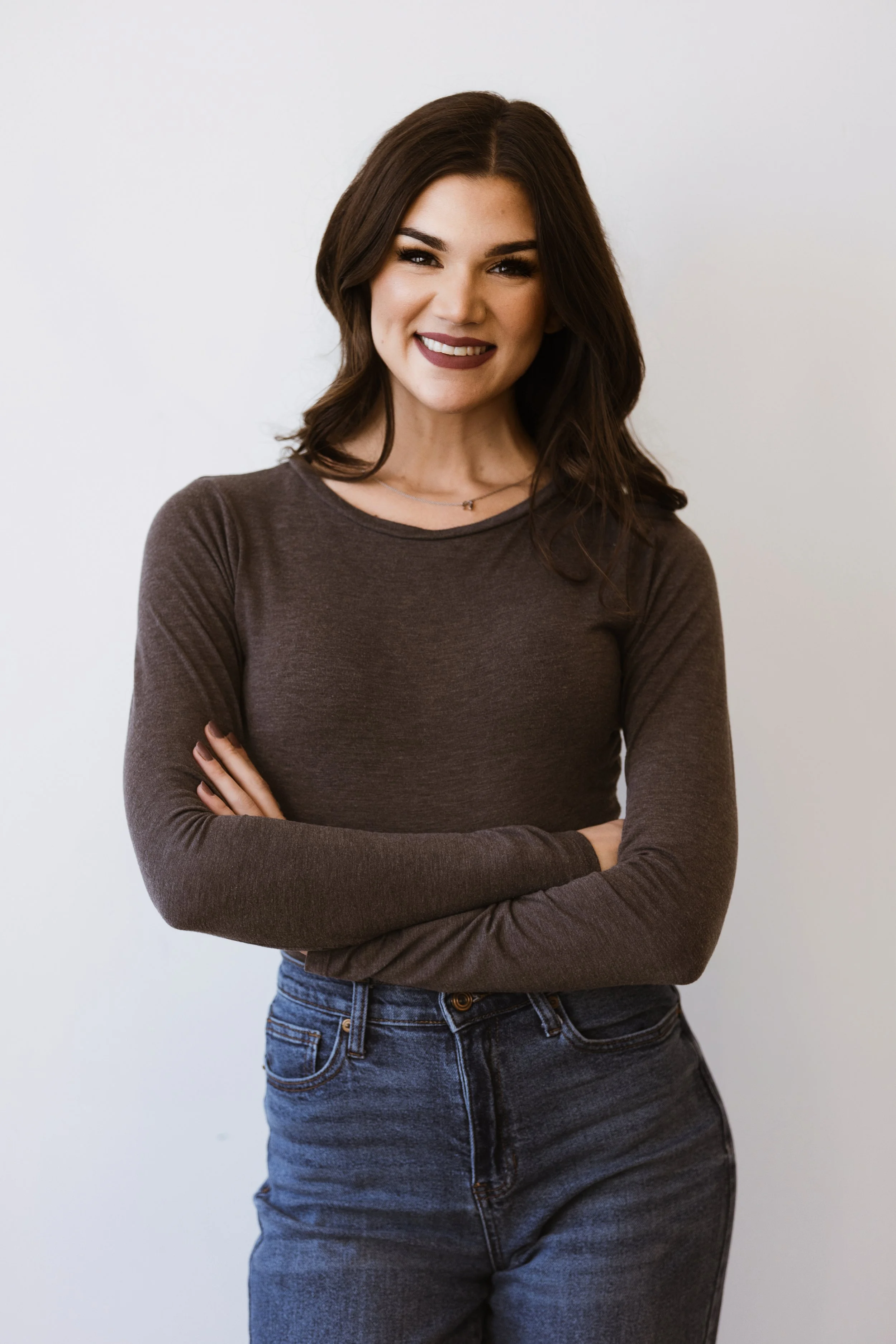A young woman with dark brown hair, wearing a dark brown long-sleeve shirt and blue jeans, smiling and standing against a plain white background.