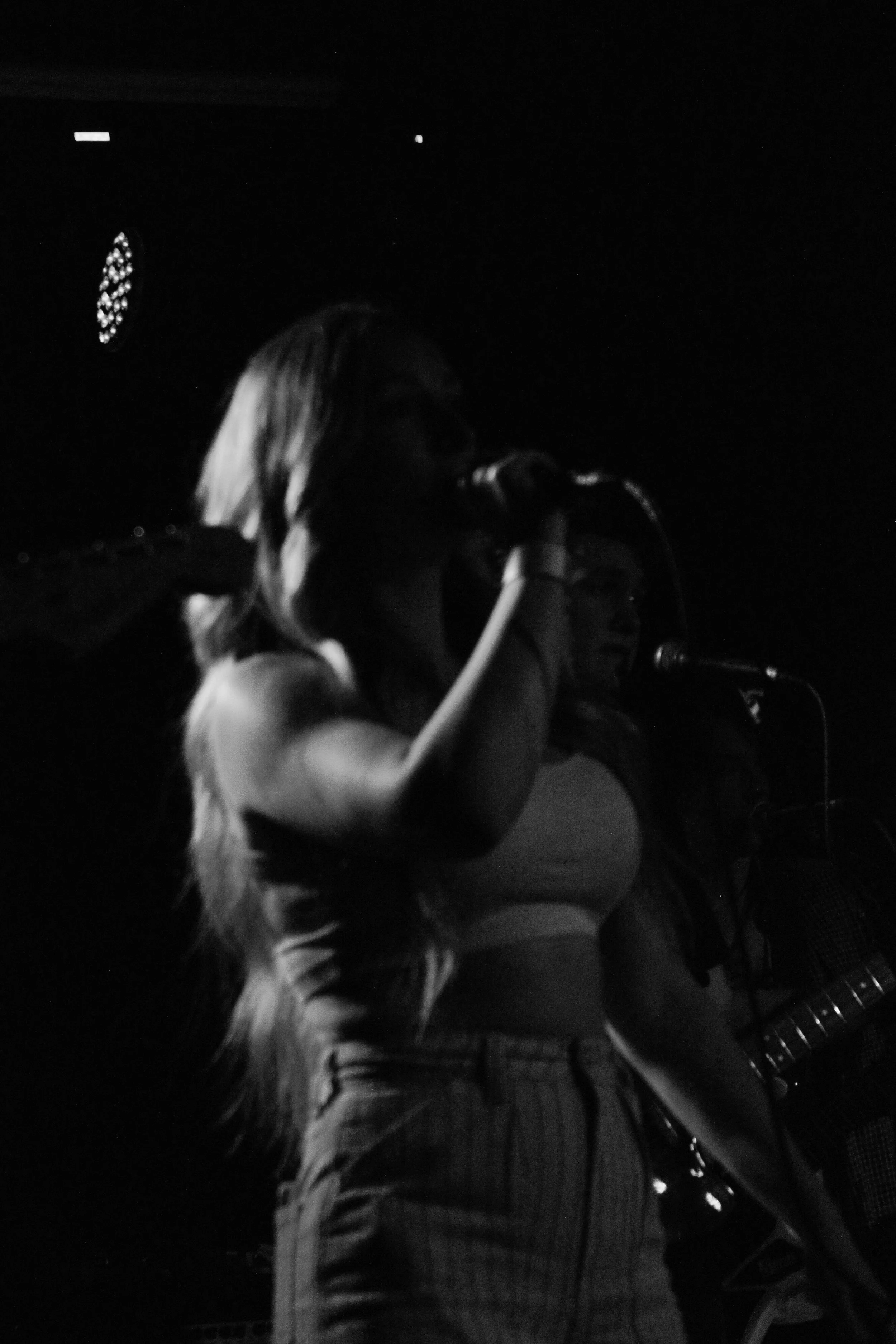 A woman with long hair wearing a sleeveless top and striped pants singing into a microphone in a dark setting.