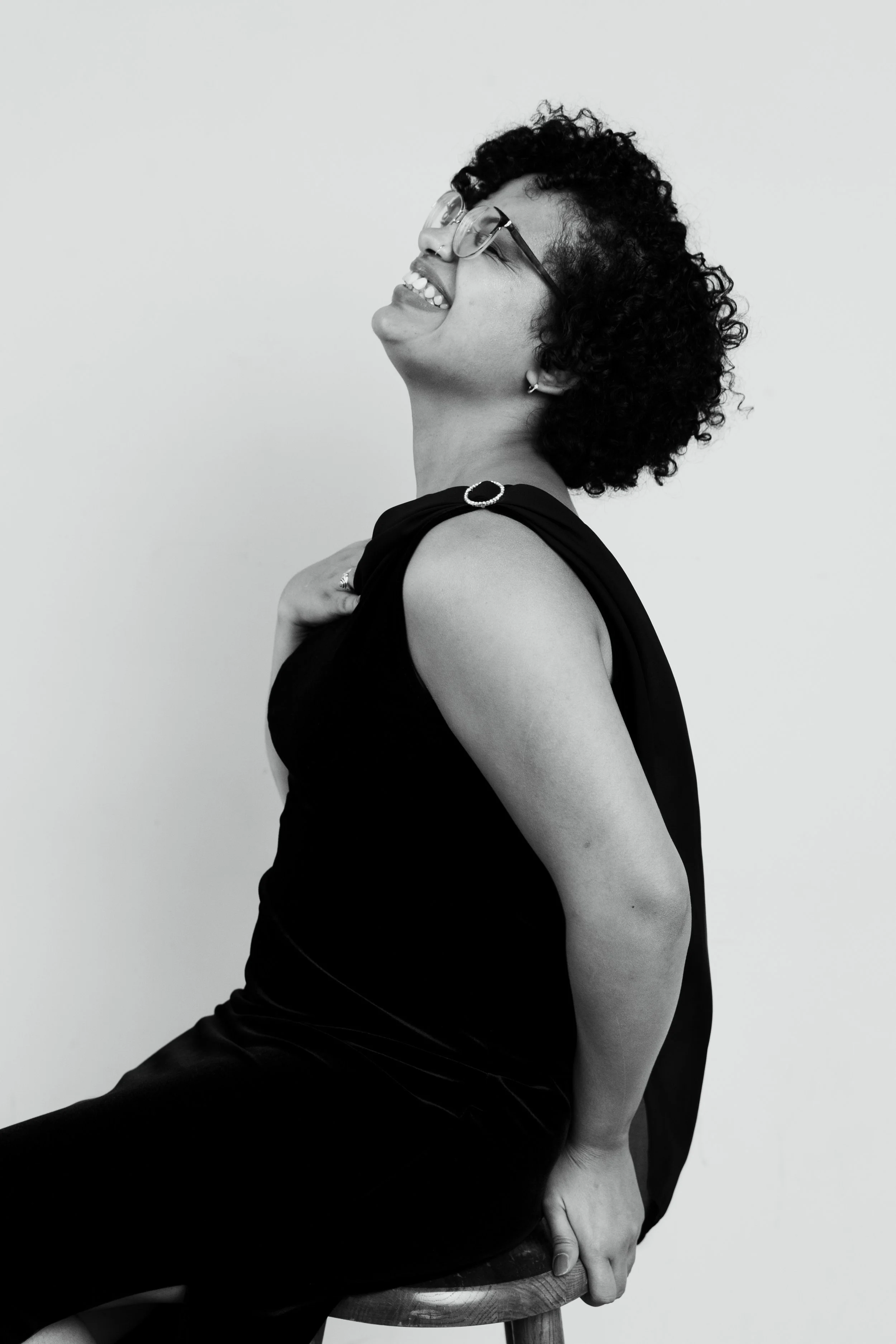 Black and white photo of a woman with curly hair wearing glasses, a sleeveless black top, and earrings, sitting on a wooden stool and smiling with eyes closed.