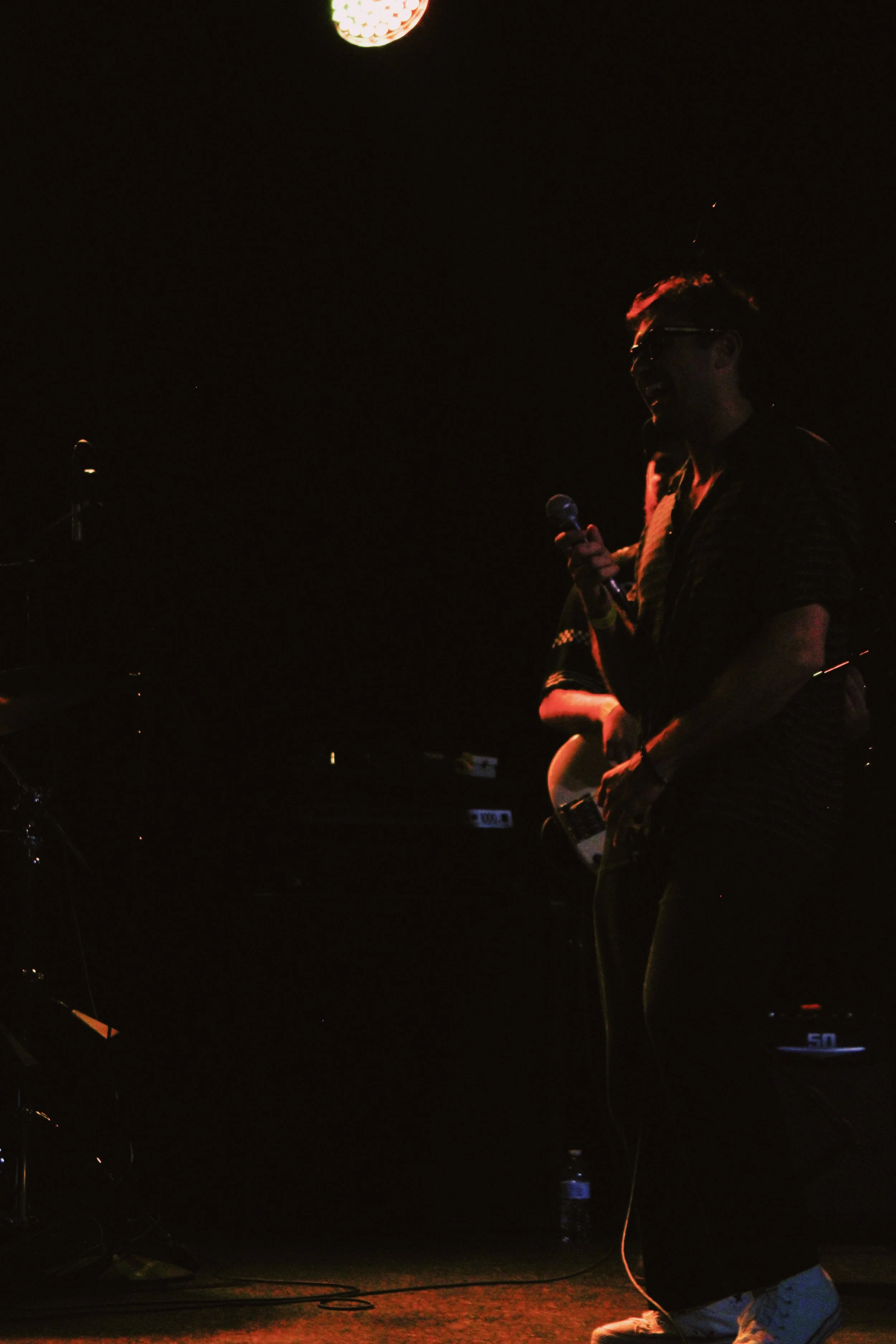 A person holding a microphone is performing on stage, silhouette against a dark background with a bright stage light overhead.