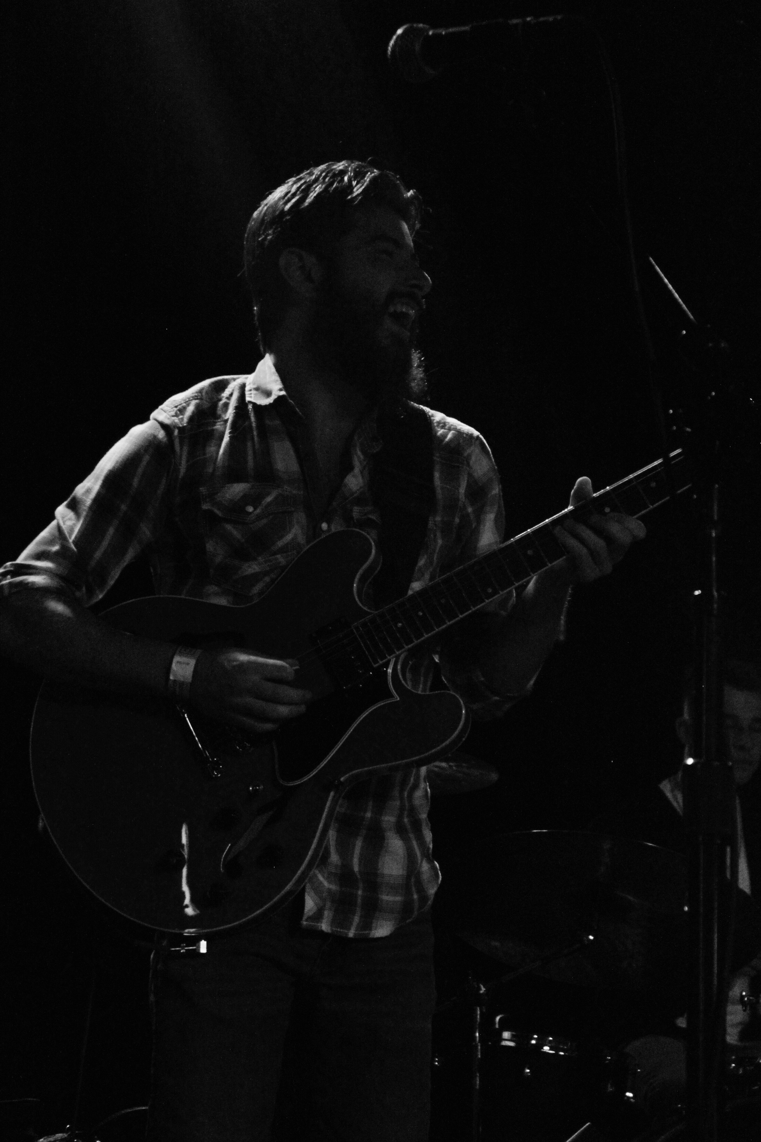 A man with a beard and plaid shirt playing an electric guitar on stage, smiling, under dim lighting.