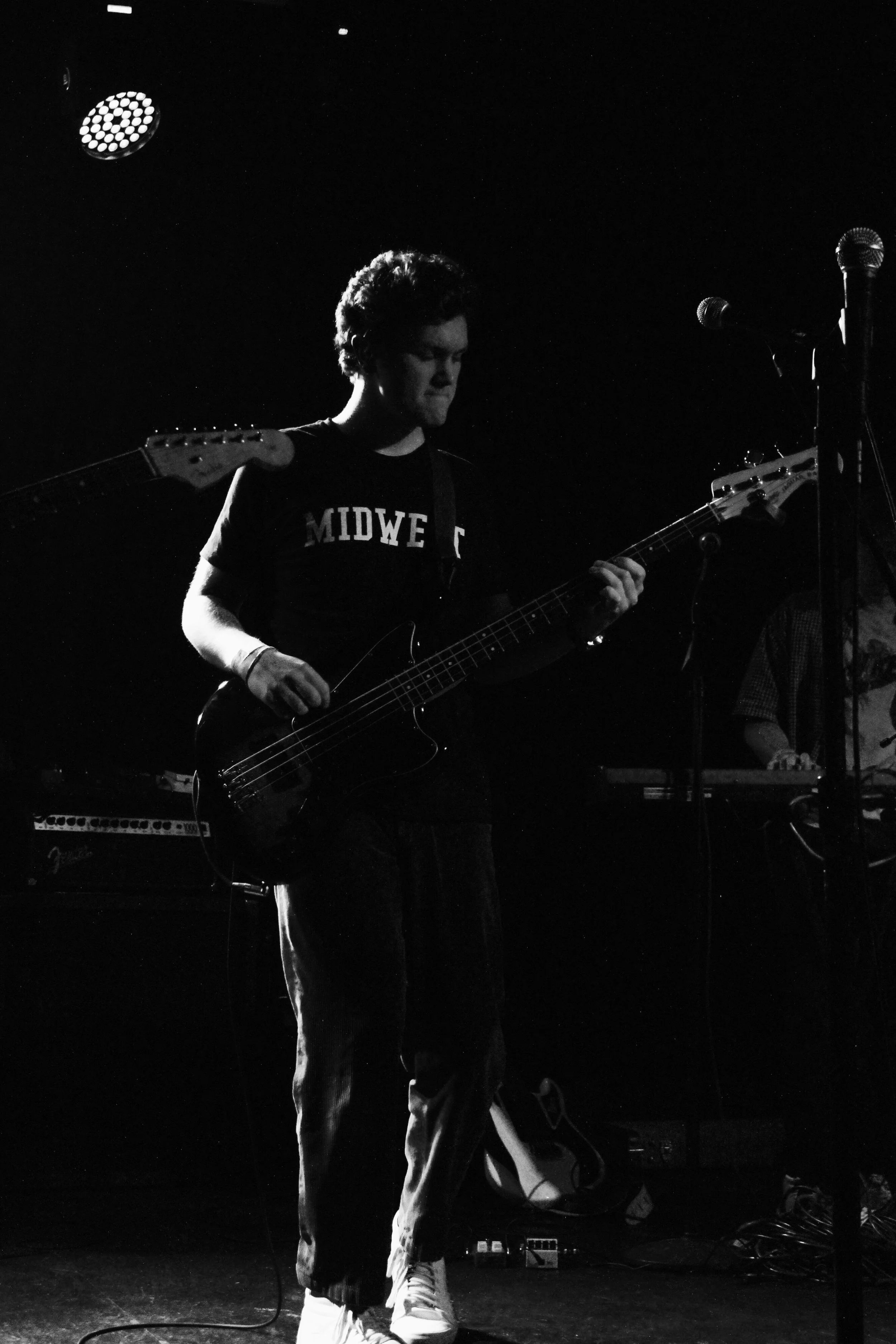 A young man playing an electric bass guitar on stage, with a keyboard player in the background, under stage lighting in a dark setting.