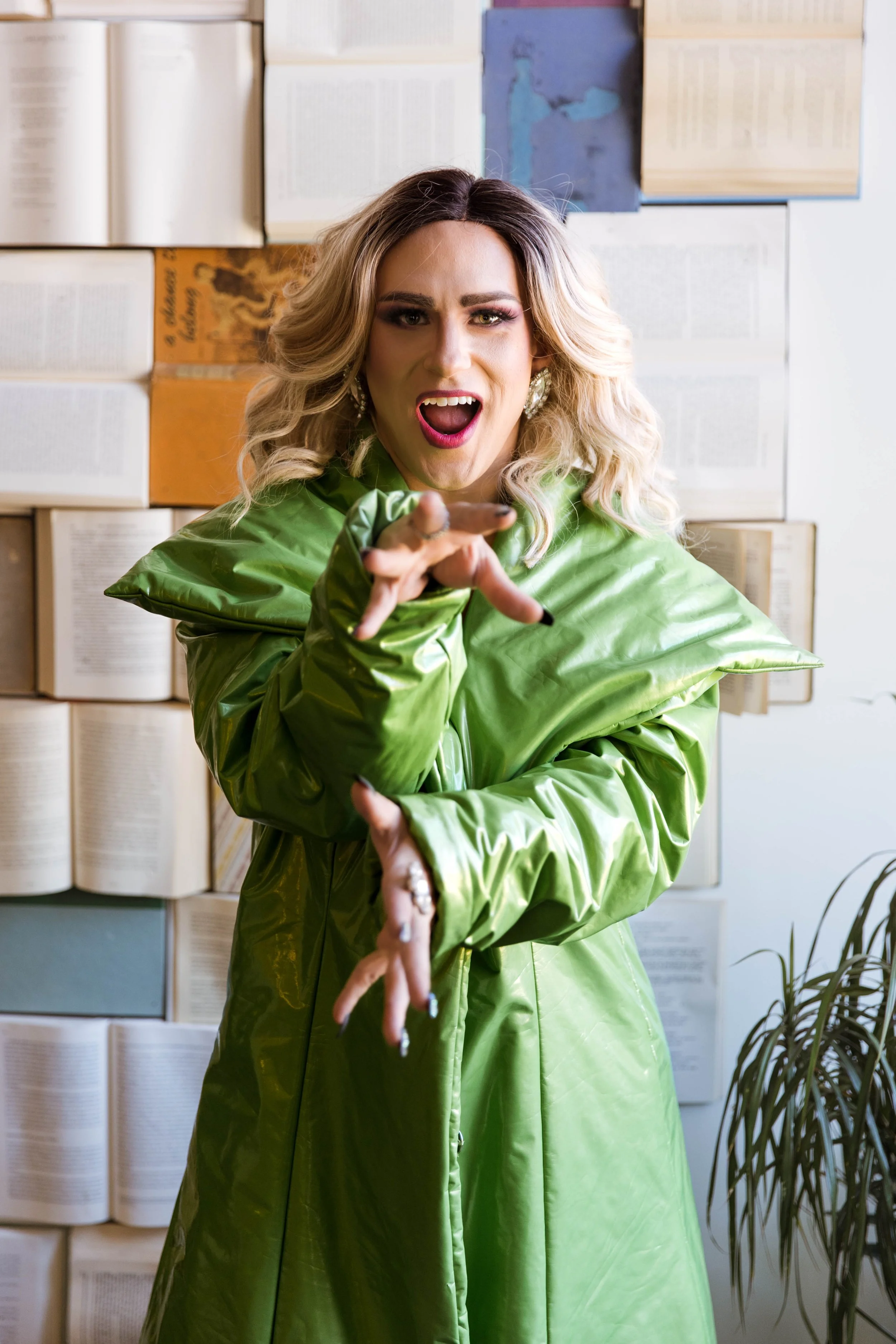 A woman with blonde curly hair, wearing a shiny green coat, gestures towards the camera with an expressive face, set against a background of open books on a wall and a potted plant.