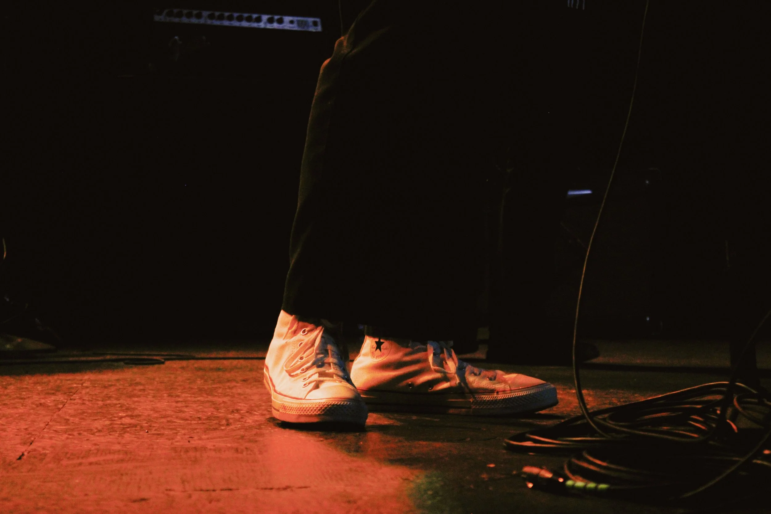 Close-up of a person's legs and feet wearing white high-top Converse sneakers, standing on a dark stage with red lighting, with audio cables and equipment nearby.