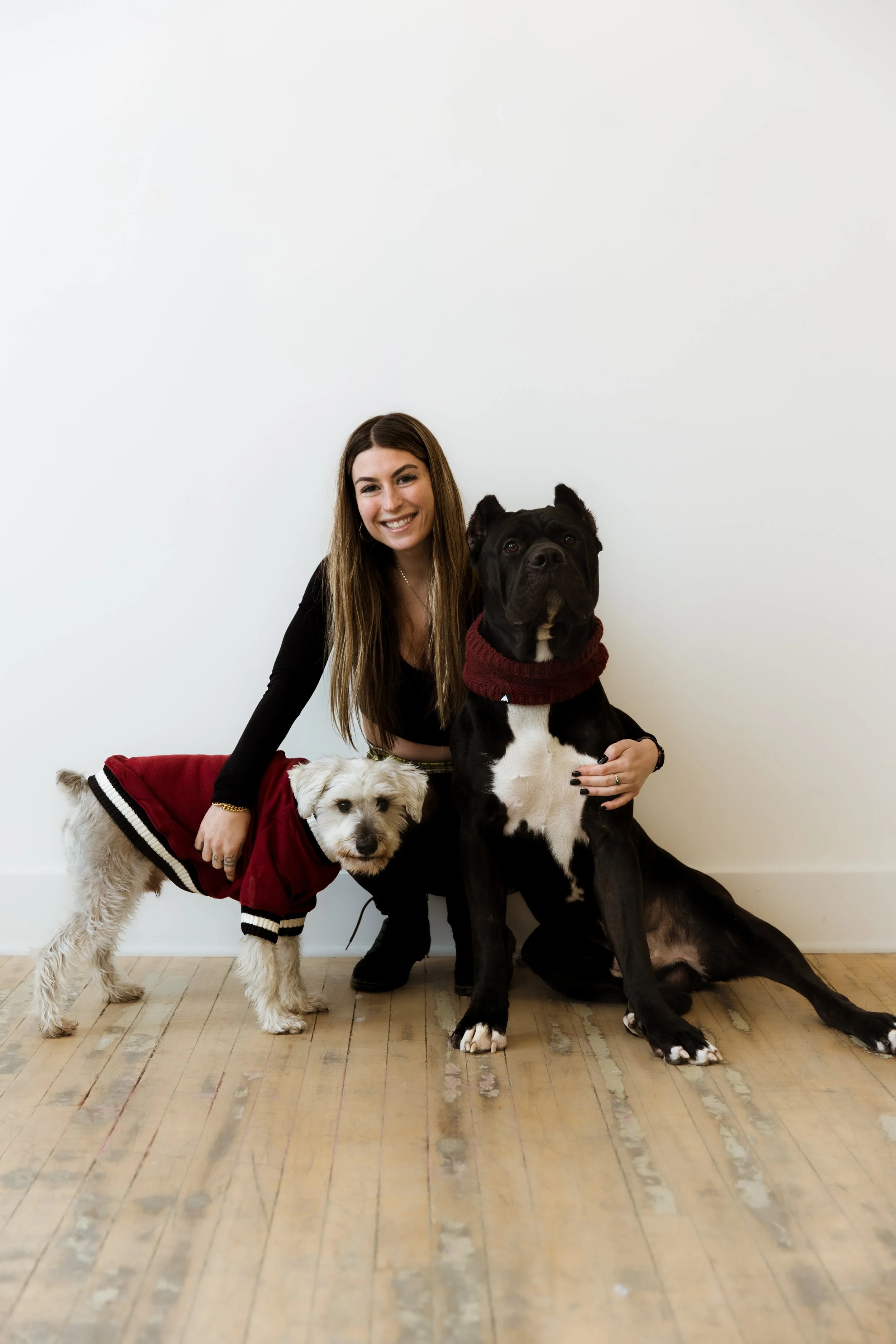 A young woman with long brown hair smiling and posing with two dogs indoors against a white wall, on a wooden floor.