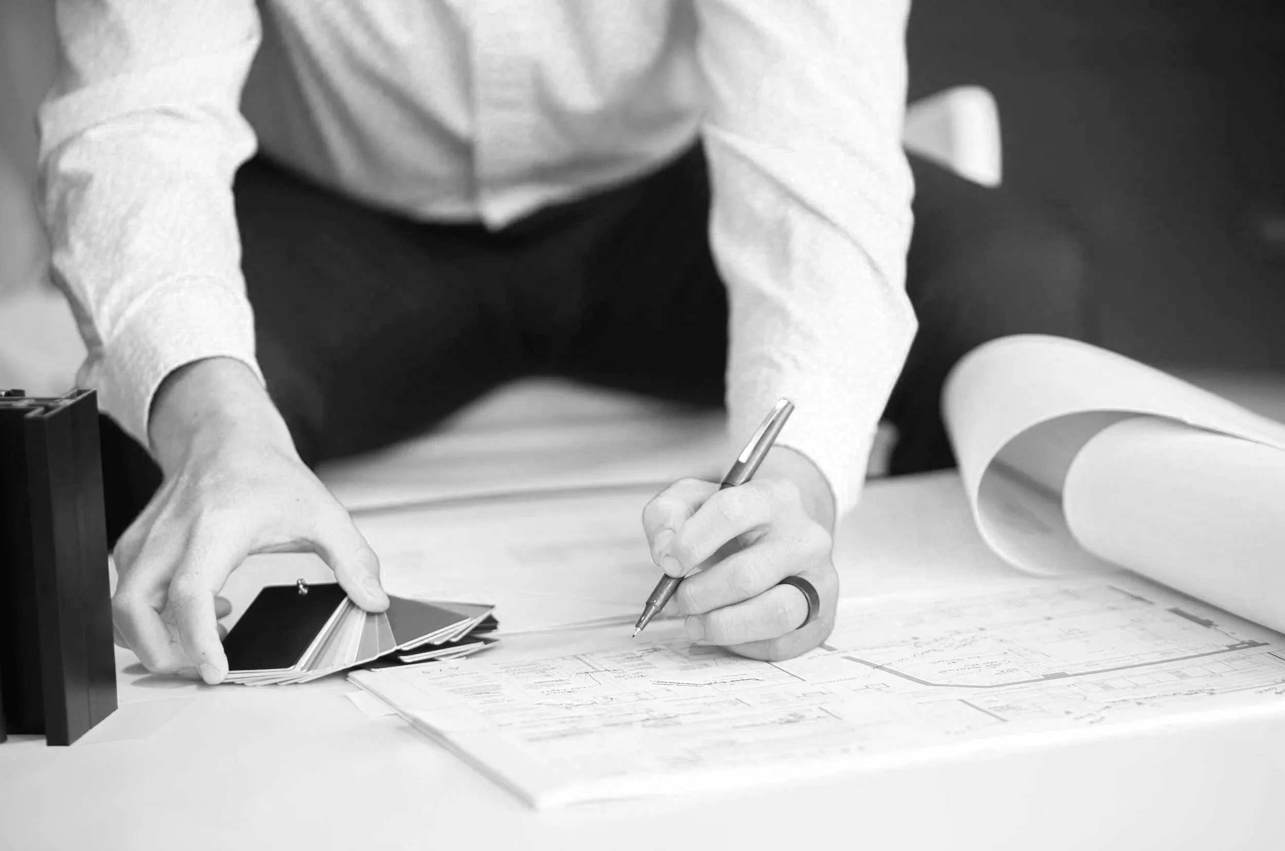 A person in a white shirt is sitting at a desk, holding a pen and writing on a blueprint or technical drawing. There are rolled-up plans and color swatches or samples on the desk.
