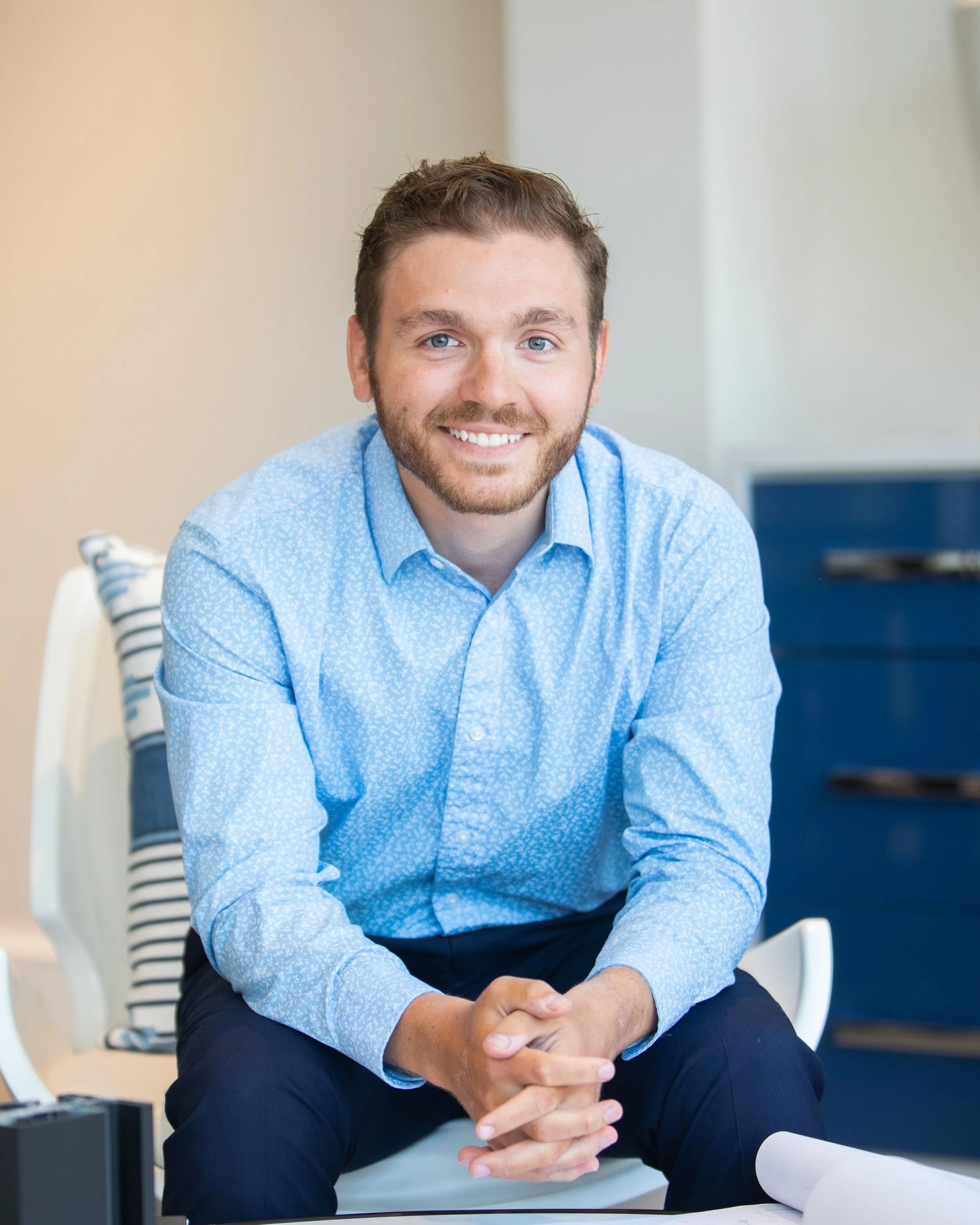 Smiling man with blue eyes and brown hair, wearing a light blue patterned shirt, sitting with hands clasped in front of him in a modern, well-lit room.