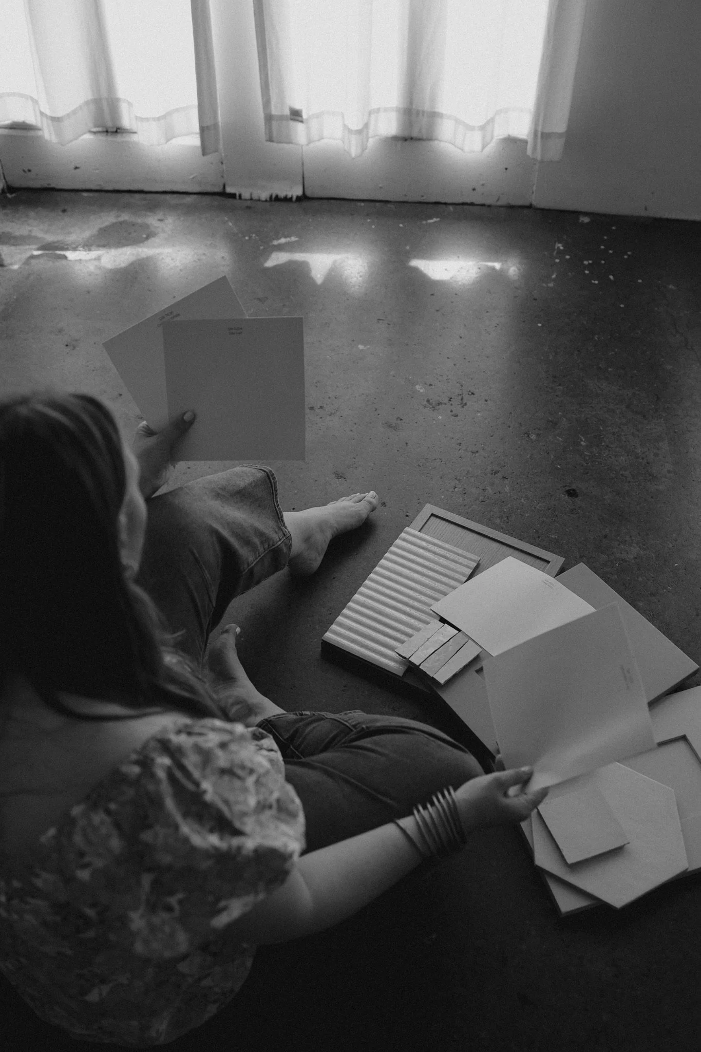 A person sitting on the floor near a window with curtains, holding papers, with more papers and empty photo frames scattered around.