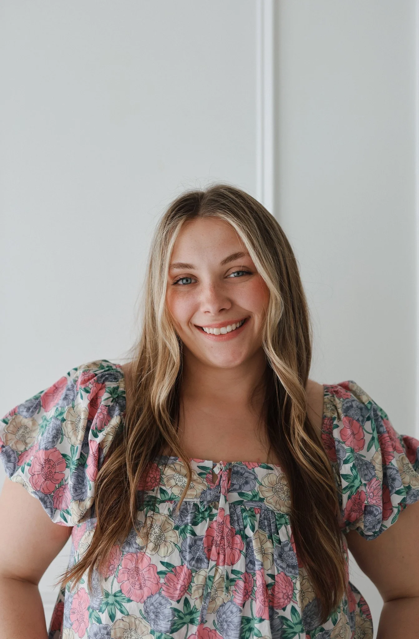 A young woman with long wavy hair smiling, wearing a floral patterned blouse with puffed sleeves, standing inside near a doorway with a white background.