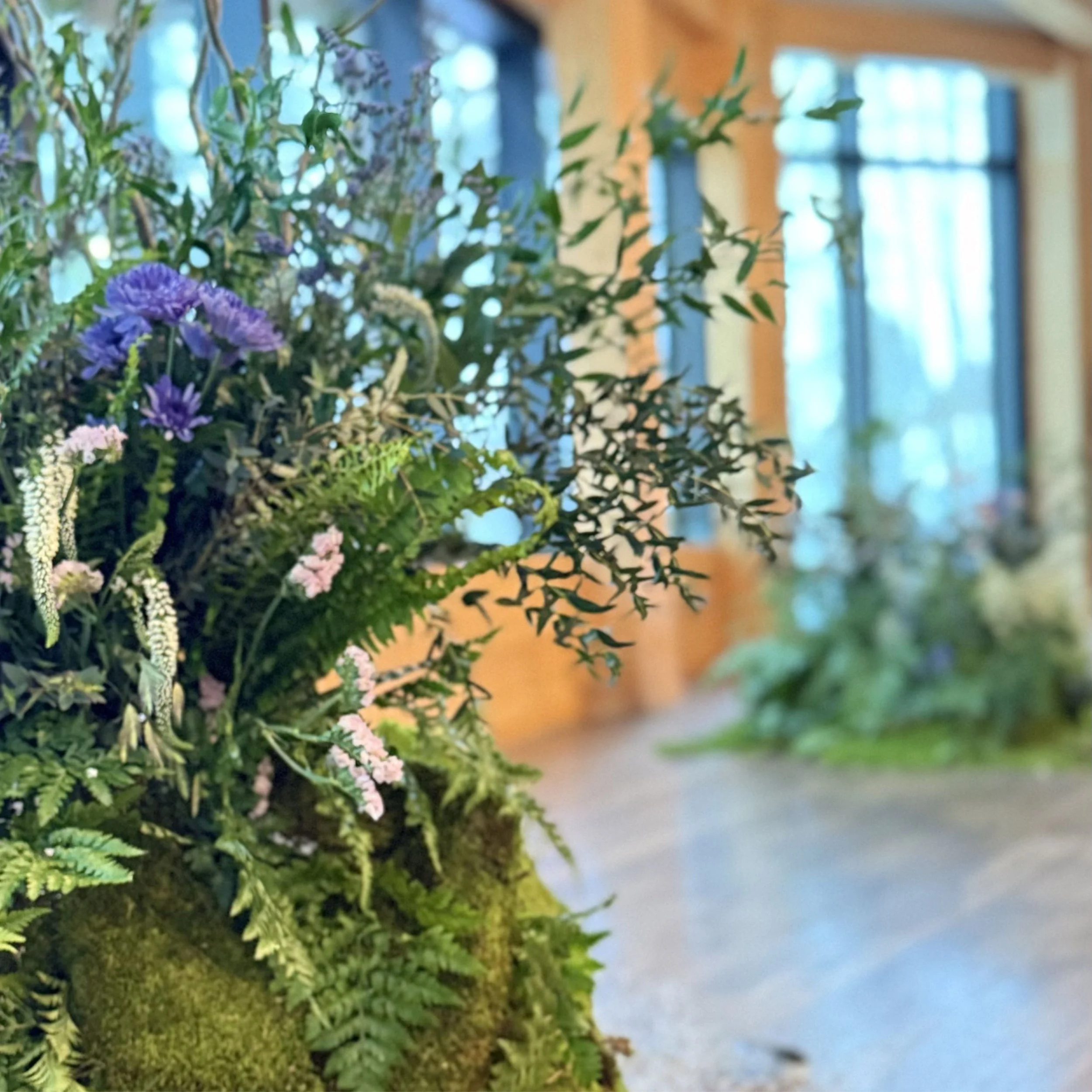 Vase de fleurs composé de différentes plantes et fleurs colorées, placé sur une surface en bois dans une pièce lumineuse avec fenêtres.