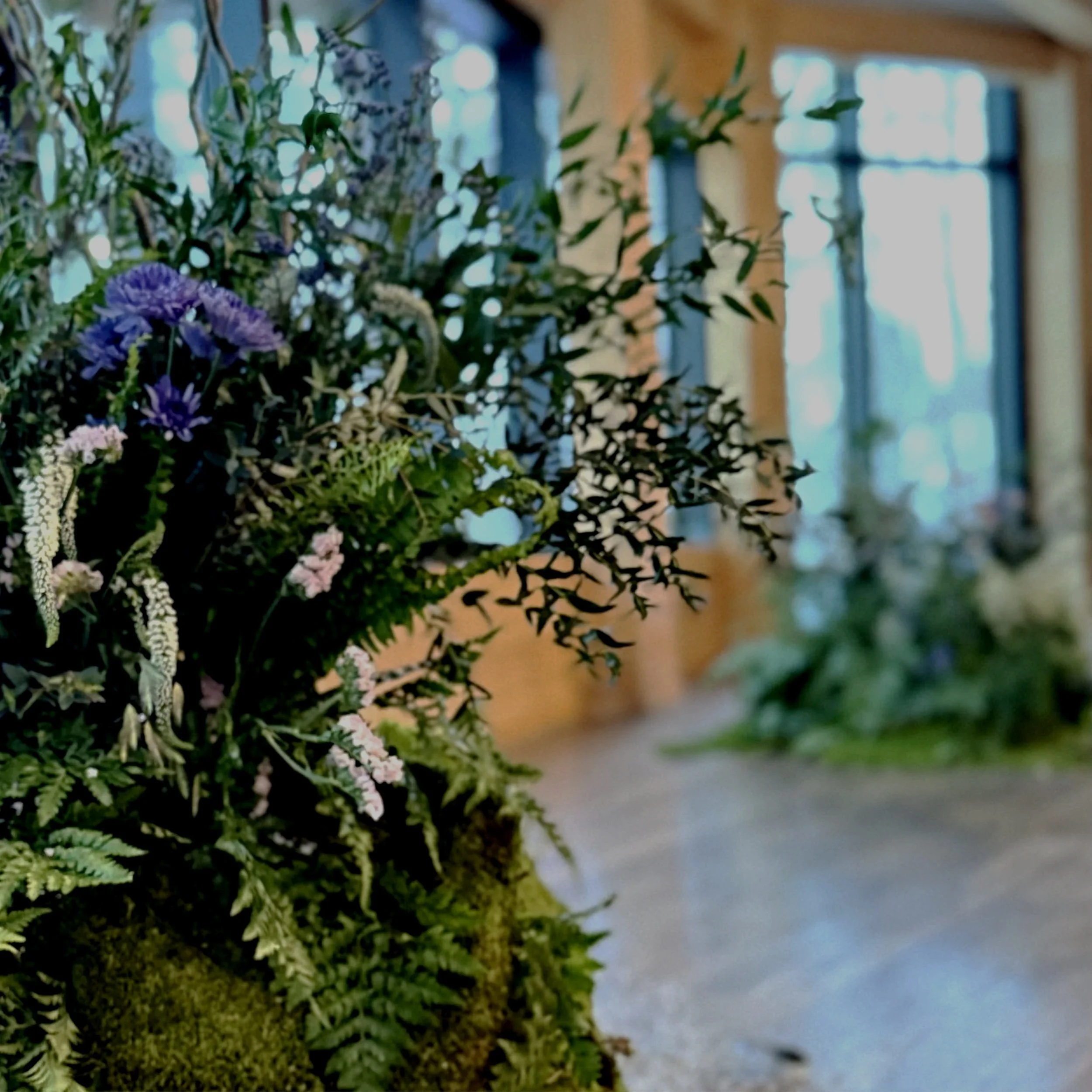 Un bouquet de fleurs composé de diverses plantes et fleurs violettes, roses, blanches et vertes, placé sur une surface en bois dans une pièce avec fenêtres en arrière-plan.