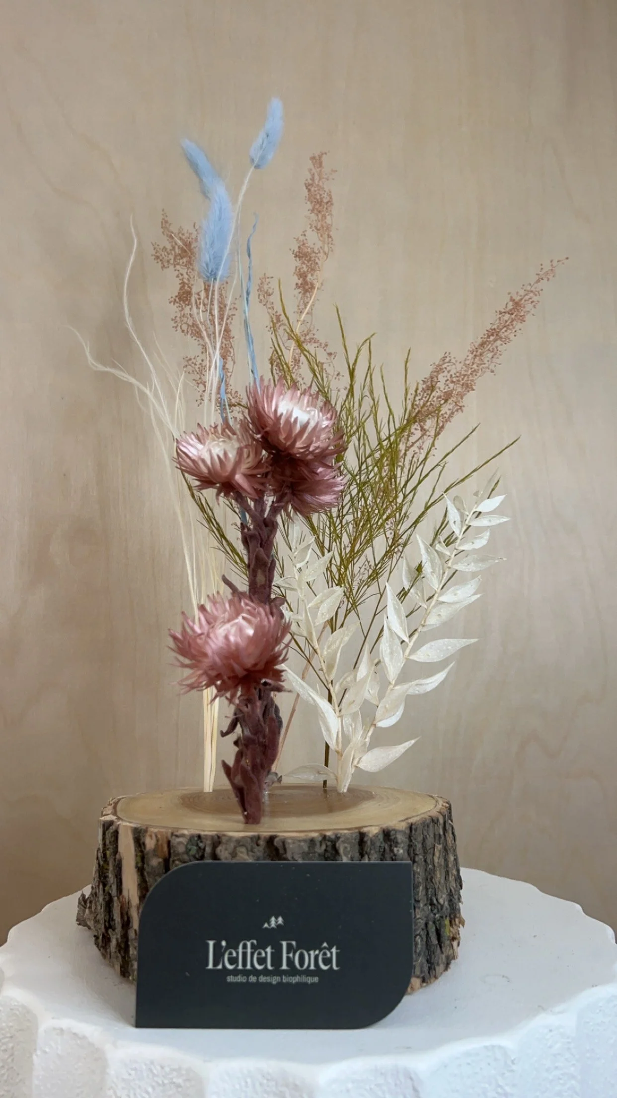 Arrangement floral avec des fleurs séchées de couleur rose, bleue, blanche et brun clair, installé sur un tronc d'arbre coupé, avec un fond neutre et un support en bois.