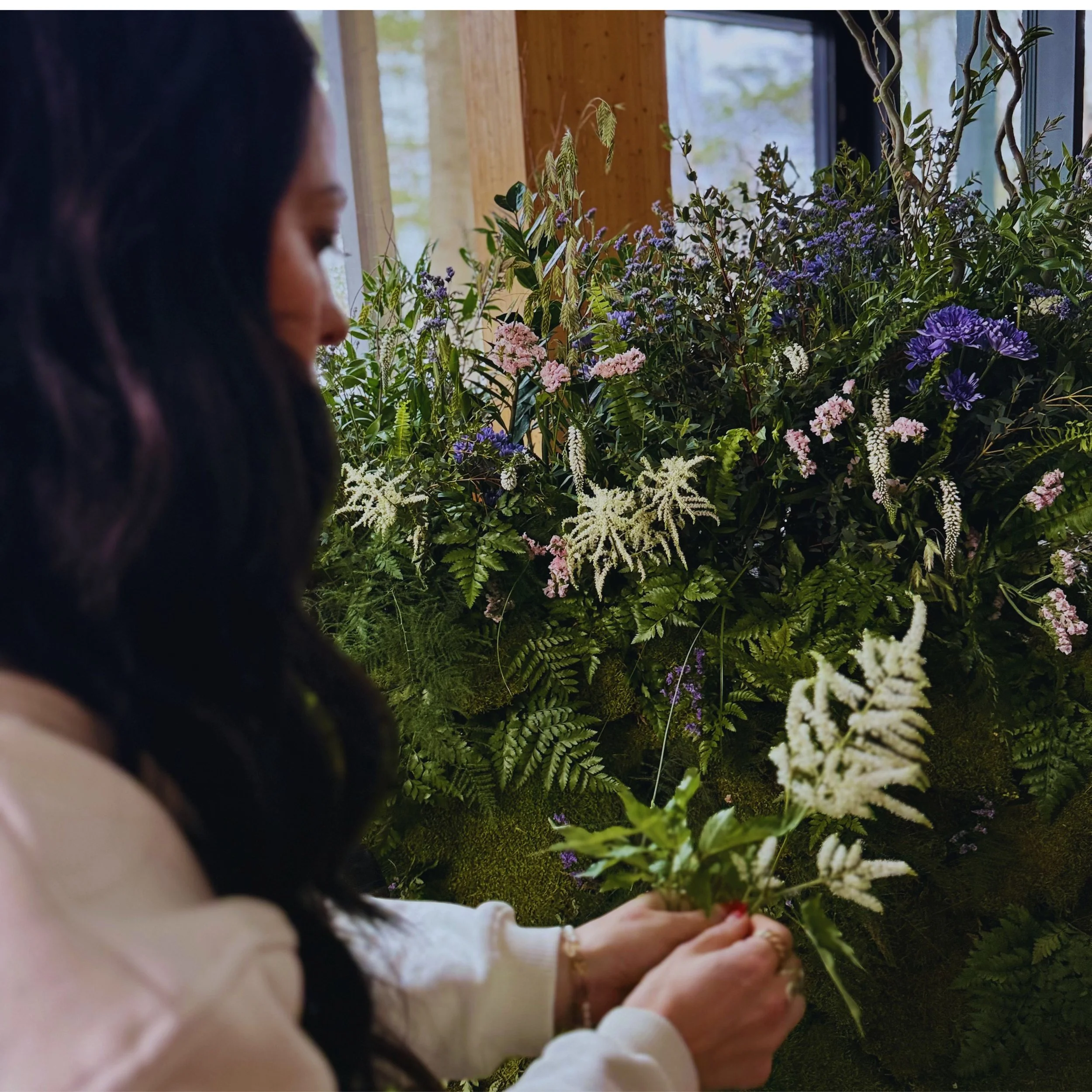 Une femme sans visage, portant un pull blanc, arrangeant ou touchant des plantes et des fleurs dans un environnement intérieur avec de grandes fenêtres laissant entrer la lumière naturelle.