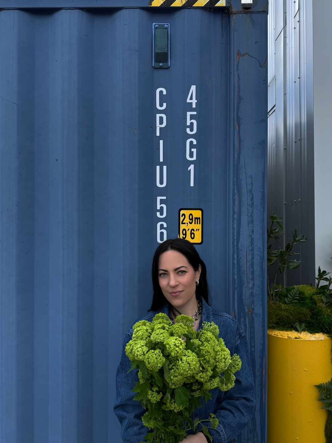 Femme tenant un bouquet de fleurs vertes devant un grand conteneur bleu avec des inscriptions et une marquage de hauteur.