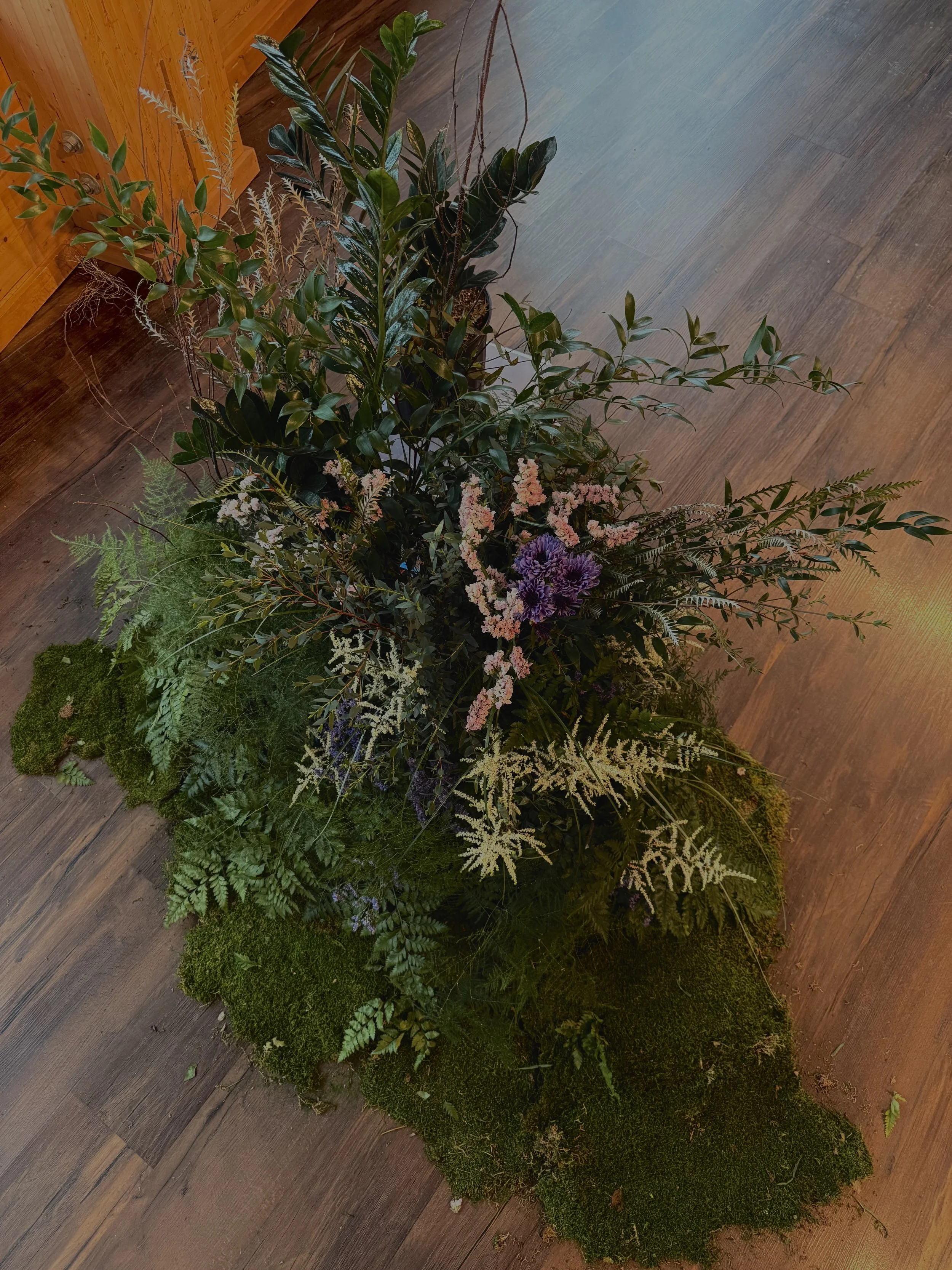 Une composition florale avec diverses plantes vertes, des fleurs roses et violettes, disposée sur un sol en bois.
