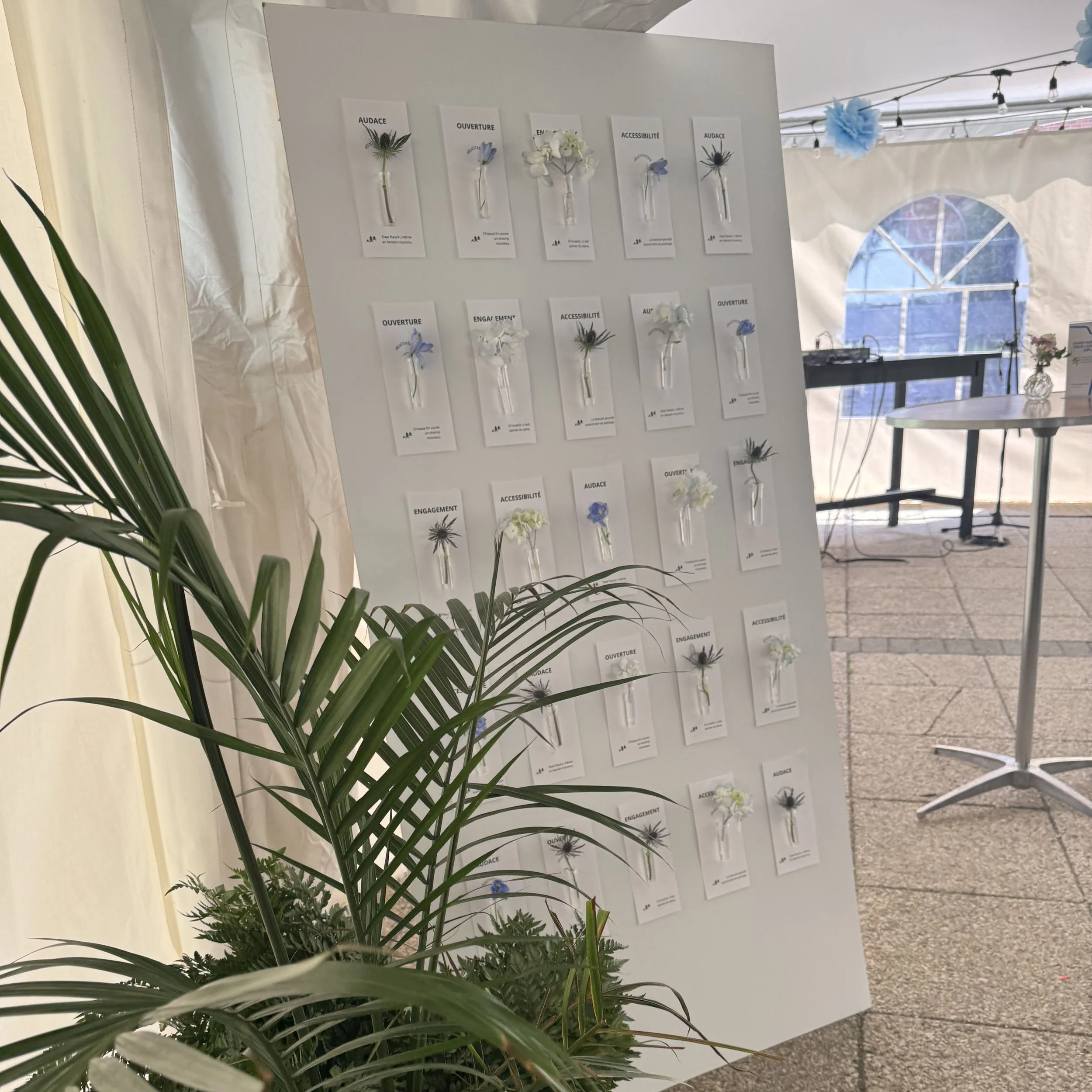 Une affiche avec des fleurs pressées en verre, chaque fleur accompagnée de mots comme 'audace', 'ouverture', 'accessibilité', et 'engagement' dans un cadre blanc, située dans un espace intérieur lumineux avec une fenêtre voisine.