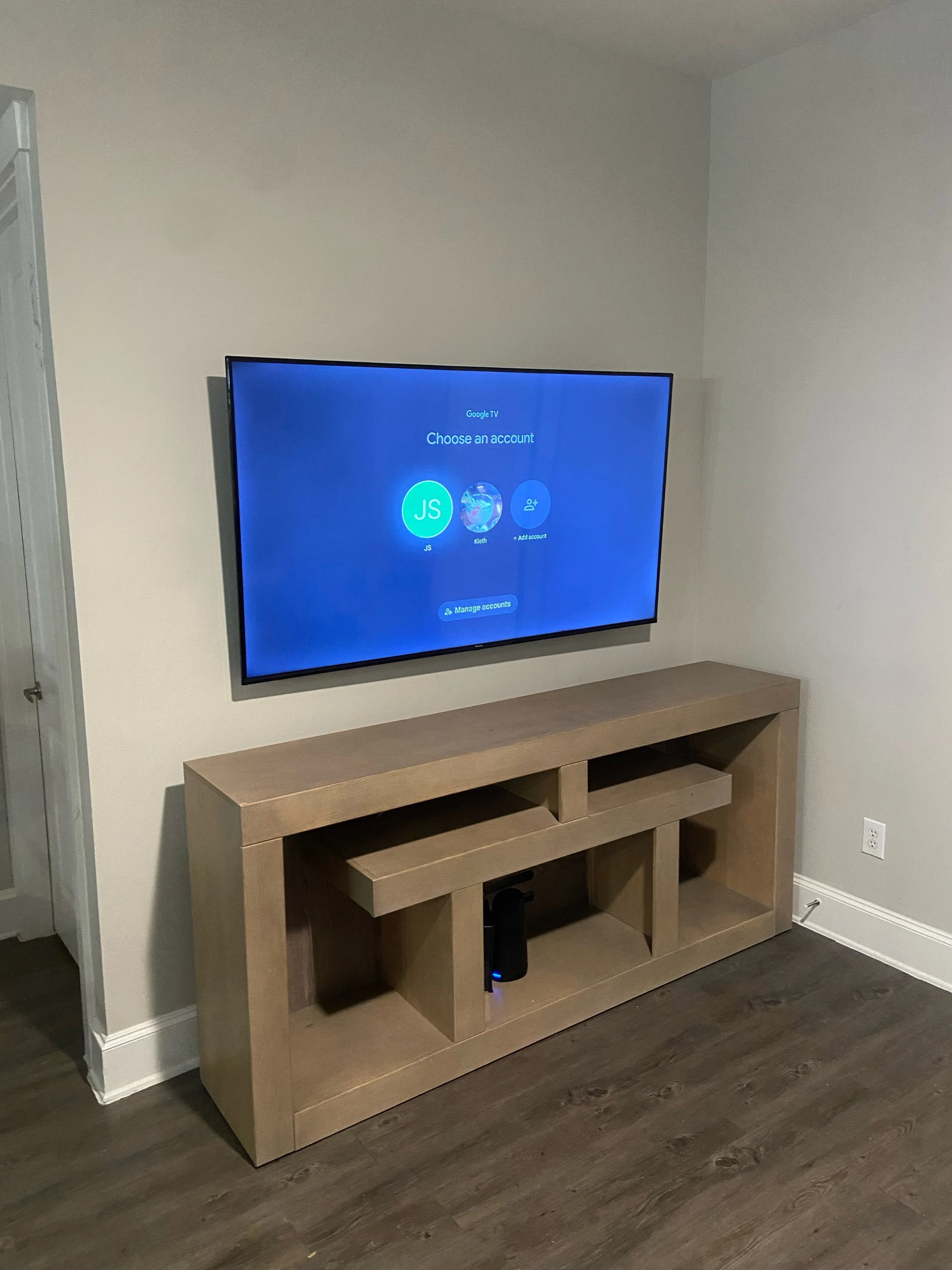 A flat-screen TV mounted on a plain white wall displaying a Google TV login screen with user profile options, positioned above a wooden console table with open shelves and a floor-standing device.