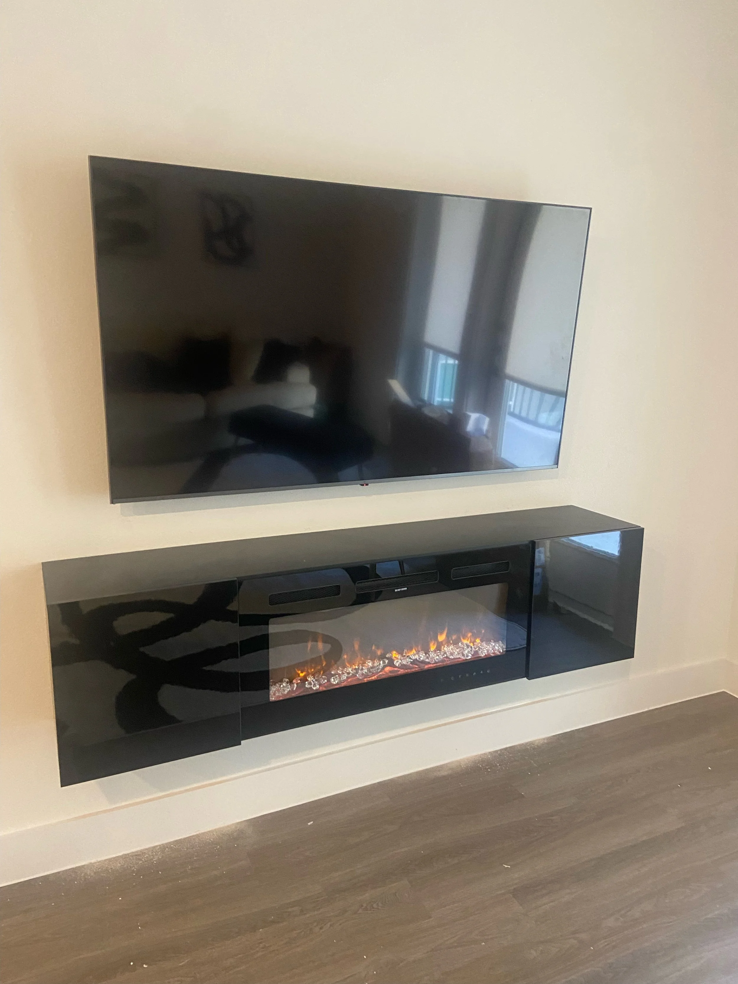 Wall-mounted flat-screen television above a black modern electric fireplace unit with a glass front and decorative crystals inside, against a beige wall with a window reflected on the TV screen.