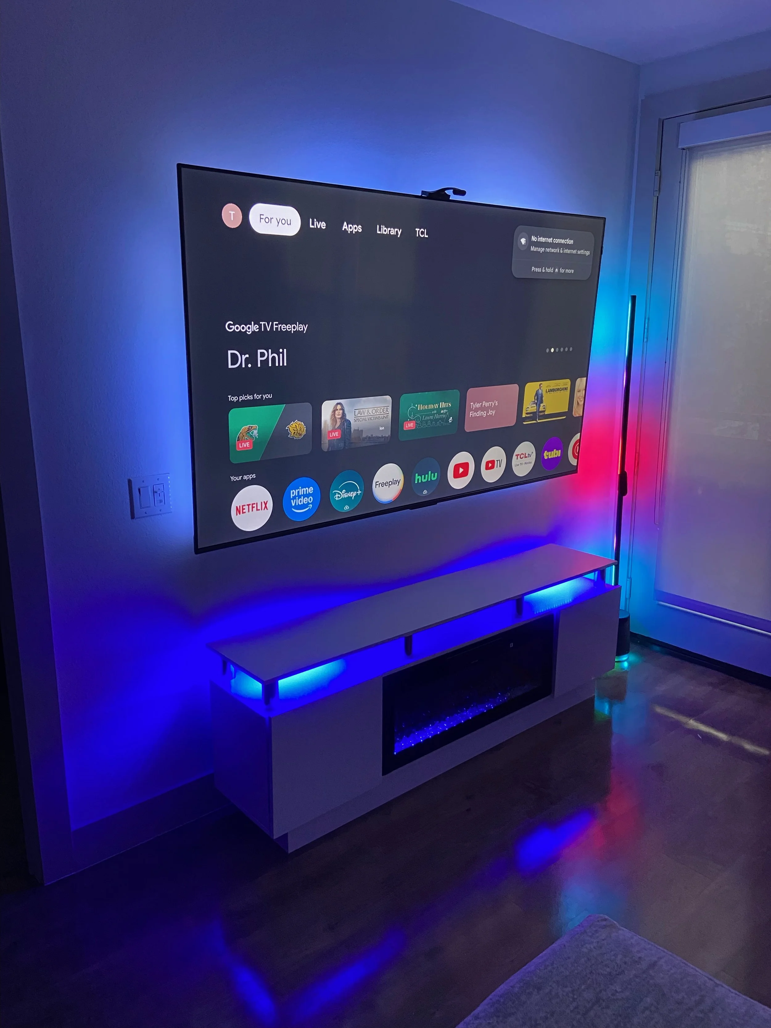 Flat-screen TV mounted on a wall displaying a streaming interface, with colorful LED lights along a white console table and a corner of a couch visible in a dimly lit room.