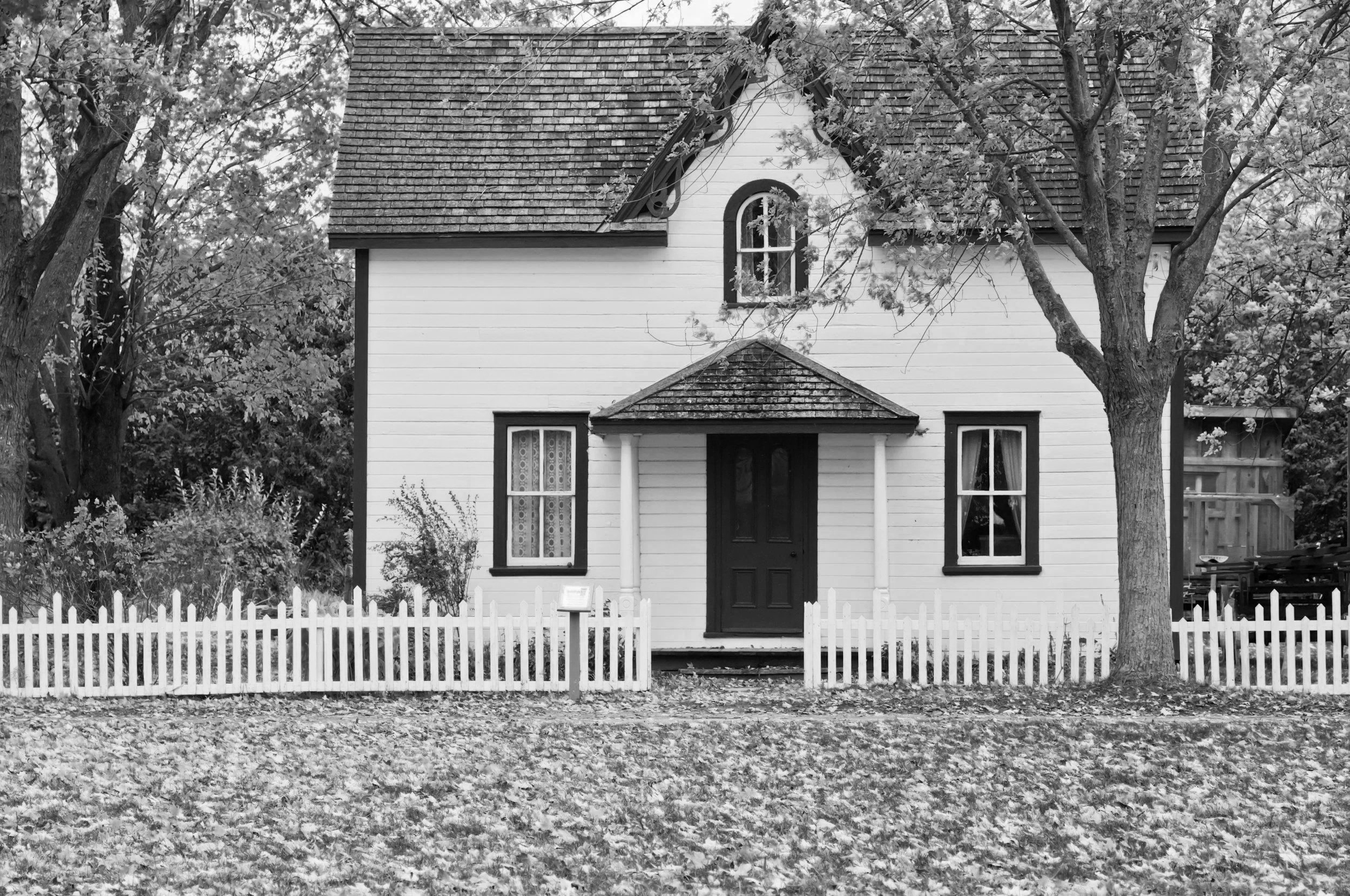 A black and white photo of a small, two-story house with a white exterior, a central front door, surrounded by trees, and enclosed by a white picket fence.