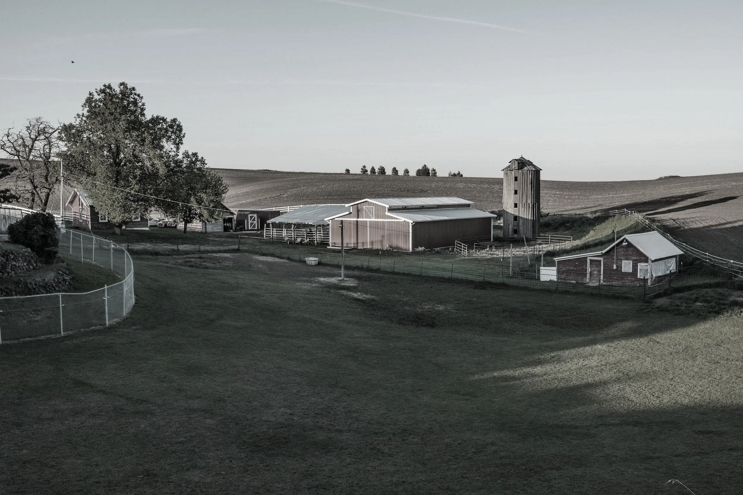 A rural farm scene with multiple barns, a silo, trees, and rolling fields in the background under a clear sky.