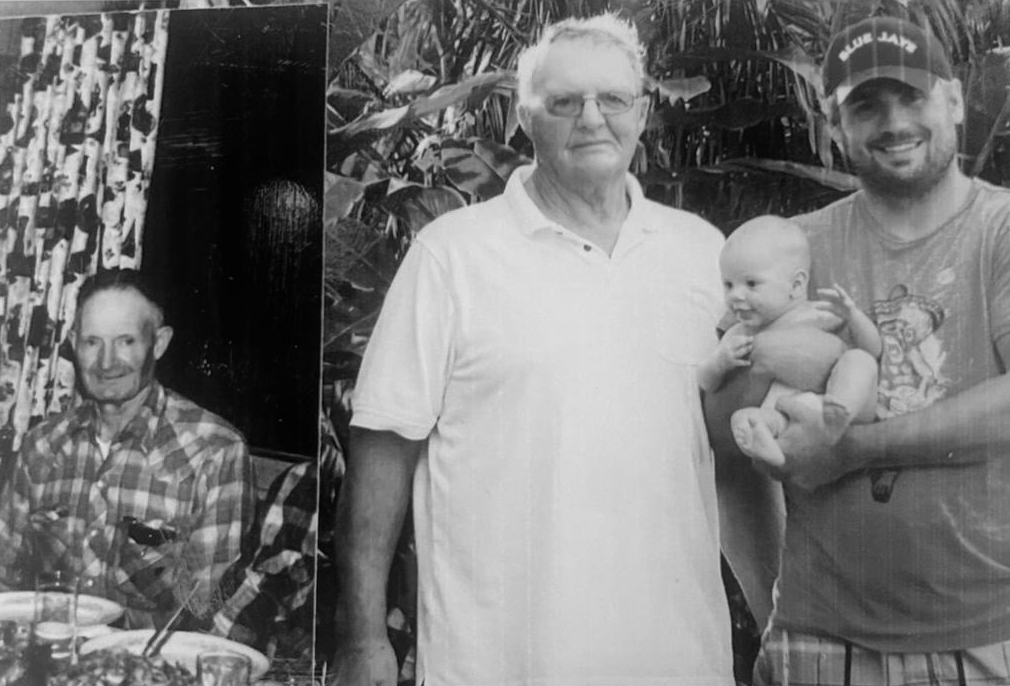 Three men and a baby outdoors, with foliage in the background. One man is older with gray hair and glasses, wearing a white shirt. The second man is younger, with a beard and cap, holding a baby. The third man is sitting indoors at a table with food, wearing a checked shirt.