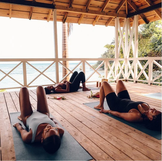Three women practicing yoga on mats on a wooden deck overlooking the beach, with palm trees and ocean in the background.