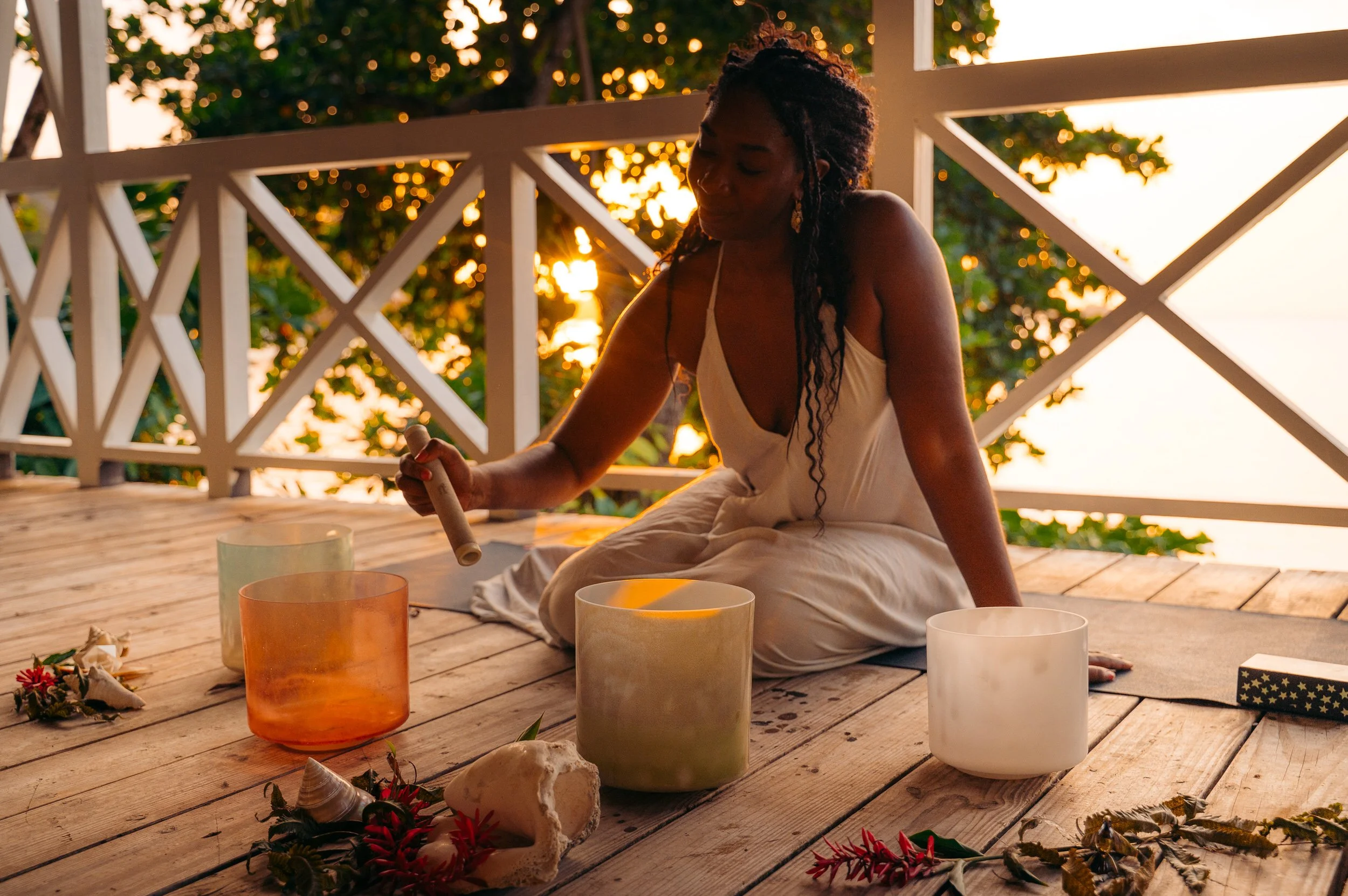 A woman in a white dress sitting cross-legged on a wooden deck during sunset, playing crystal singing bowls surrounded by flowers and seashells.