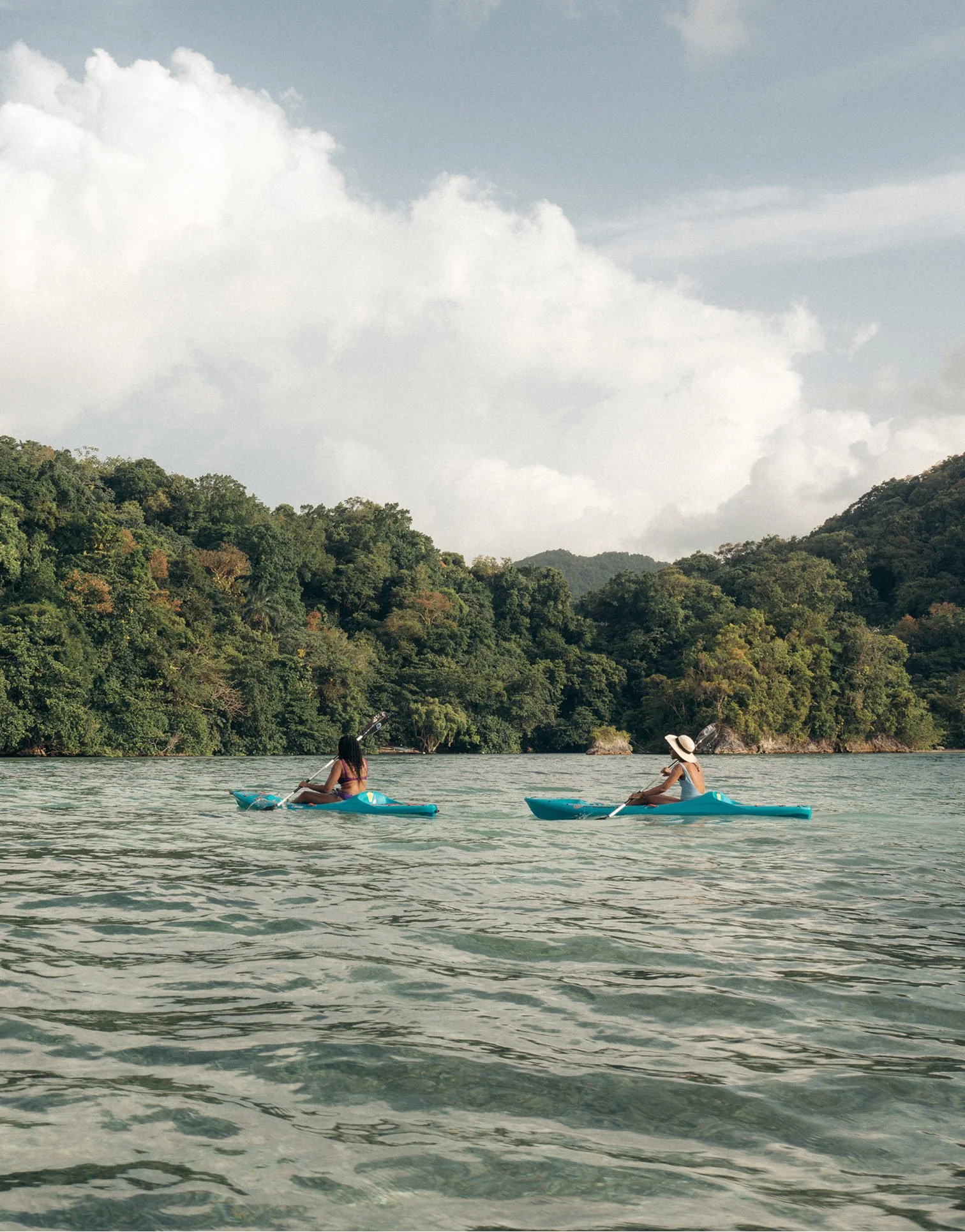 Two women kayaking on a body of water with a lush green mountainous landscape in the background, under a partly cloudy sky.