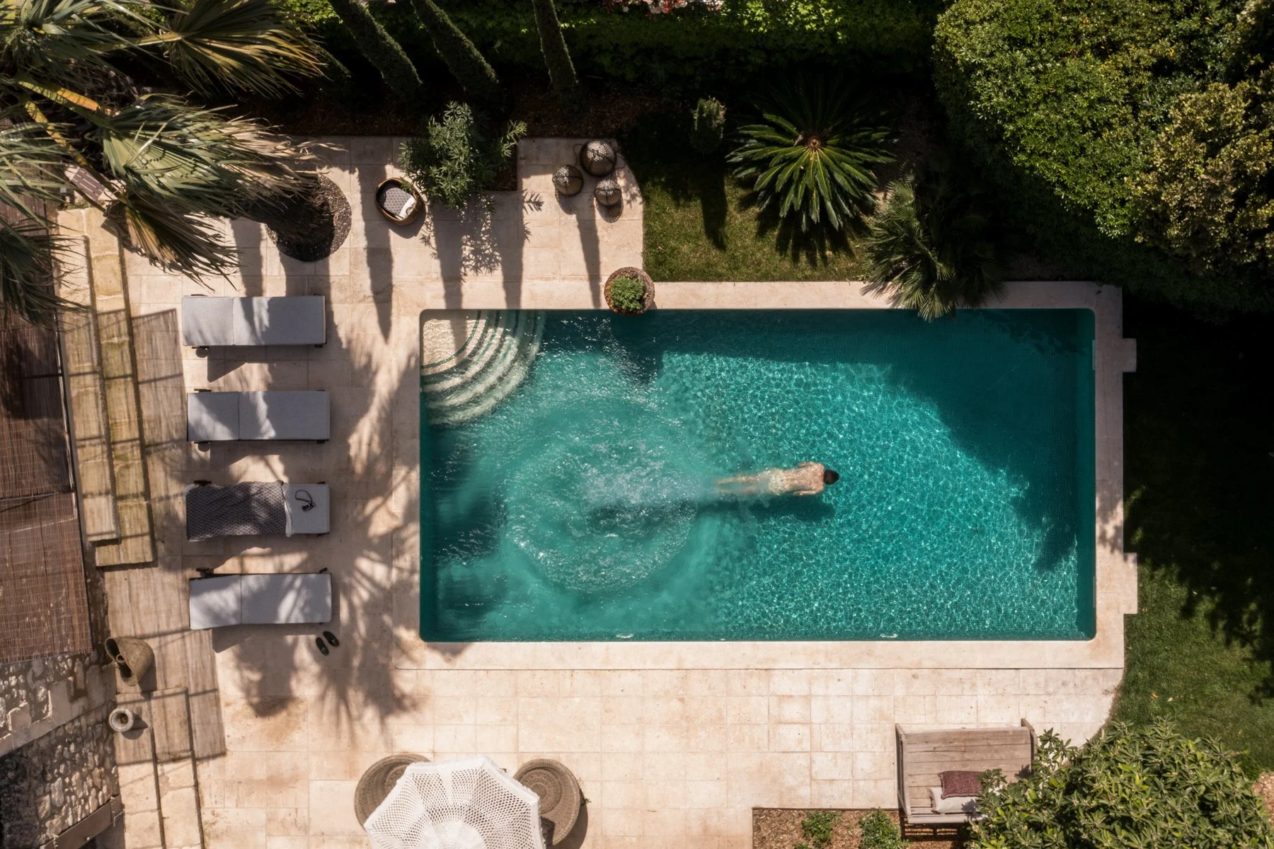 Aerial view of a backyard swimming pool with a person swimming, surrounded by a stone deck, lounge chairs, potted plants, and lush greenery.