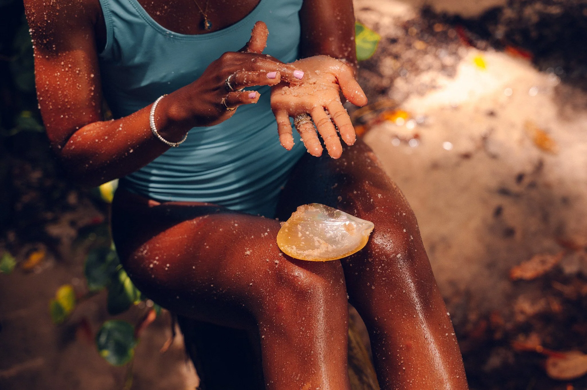 A woman with dark skin and dressed in a blue swimsuit is sitting outdoors, holding a piece of sea glass in her left hand and a handful of grainy sand in her right hand. She is seated with her legs bent and dusted with sand, with a blurred sandy and leafy background.