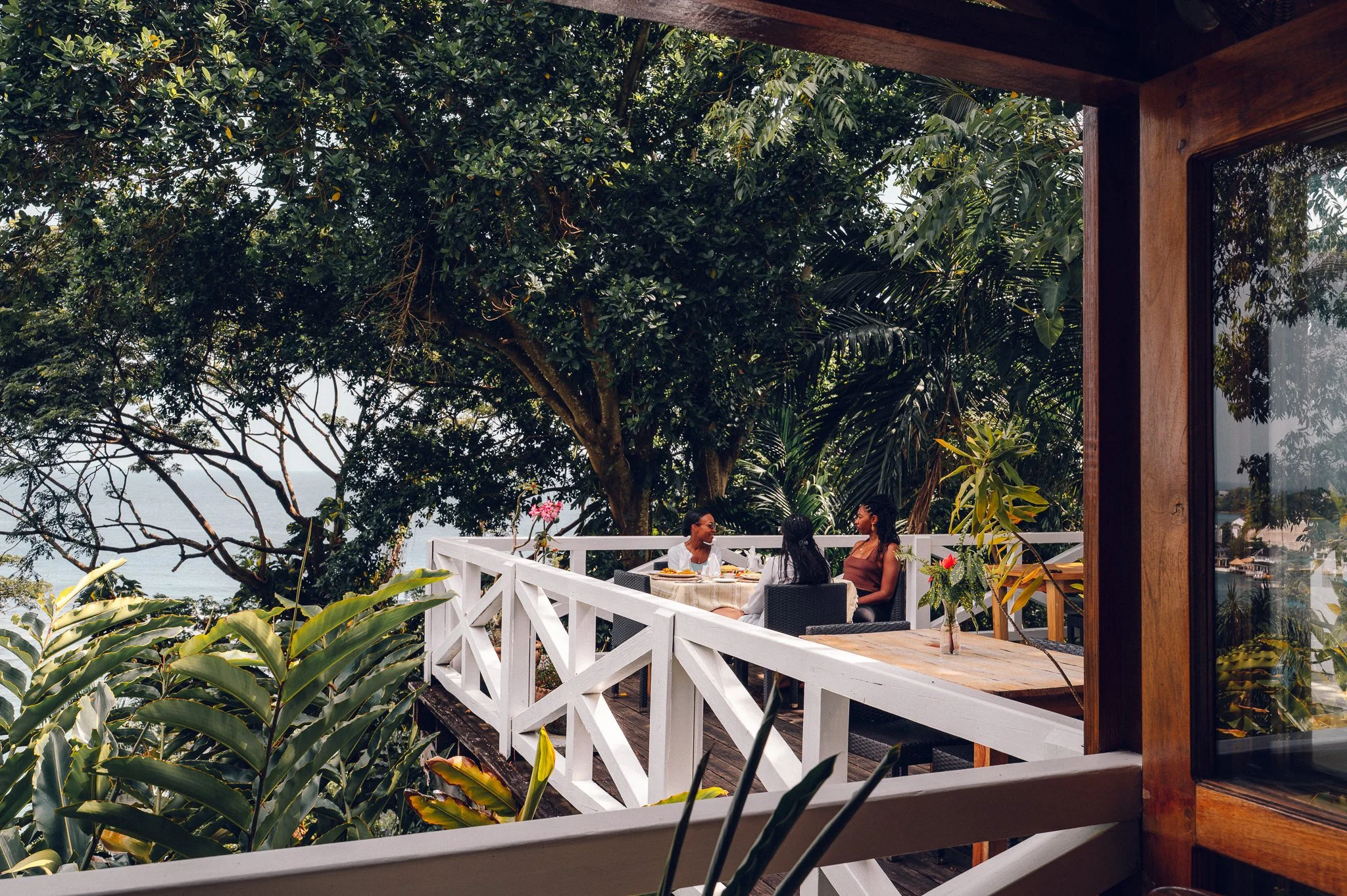 Three women sitting at a table on a balcony surrounded by lush greenery, with trees and plants, enjoying a meal or drinks.