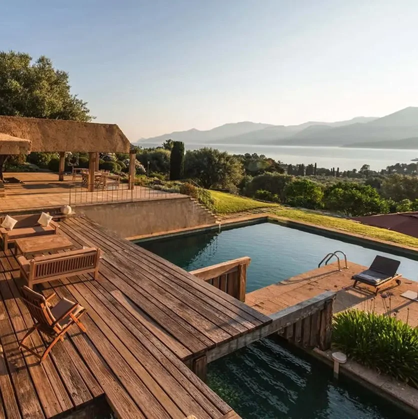 Wooden deck with outdoor furniture overlooking a swimming pool, lush greenery, and mountains in the distance under a clear sky.