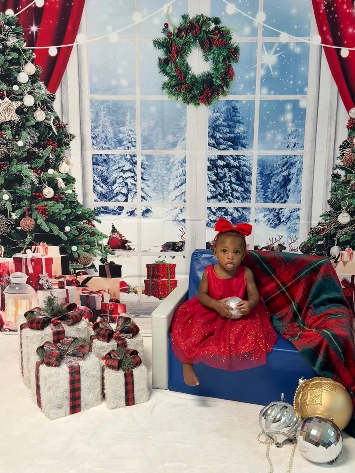 A young girl in a red dress with a large red bow on her head sitting on a blue bench surrounded by Christmas decorations. She is holding a silver ornament. Behind her is a large window showing a snowy winter scene with snow-covered trees. The scene i