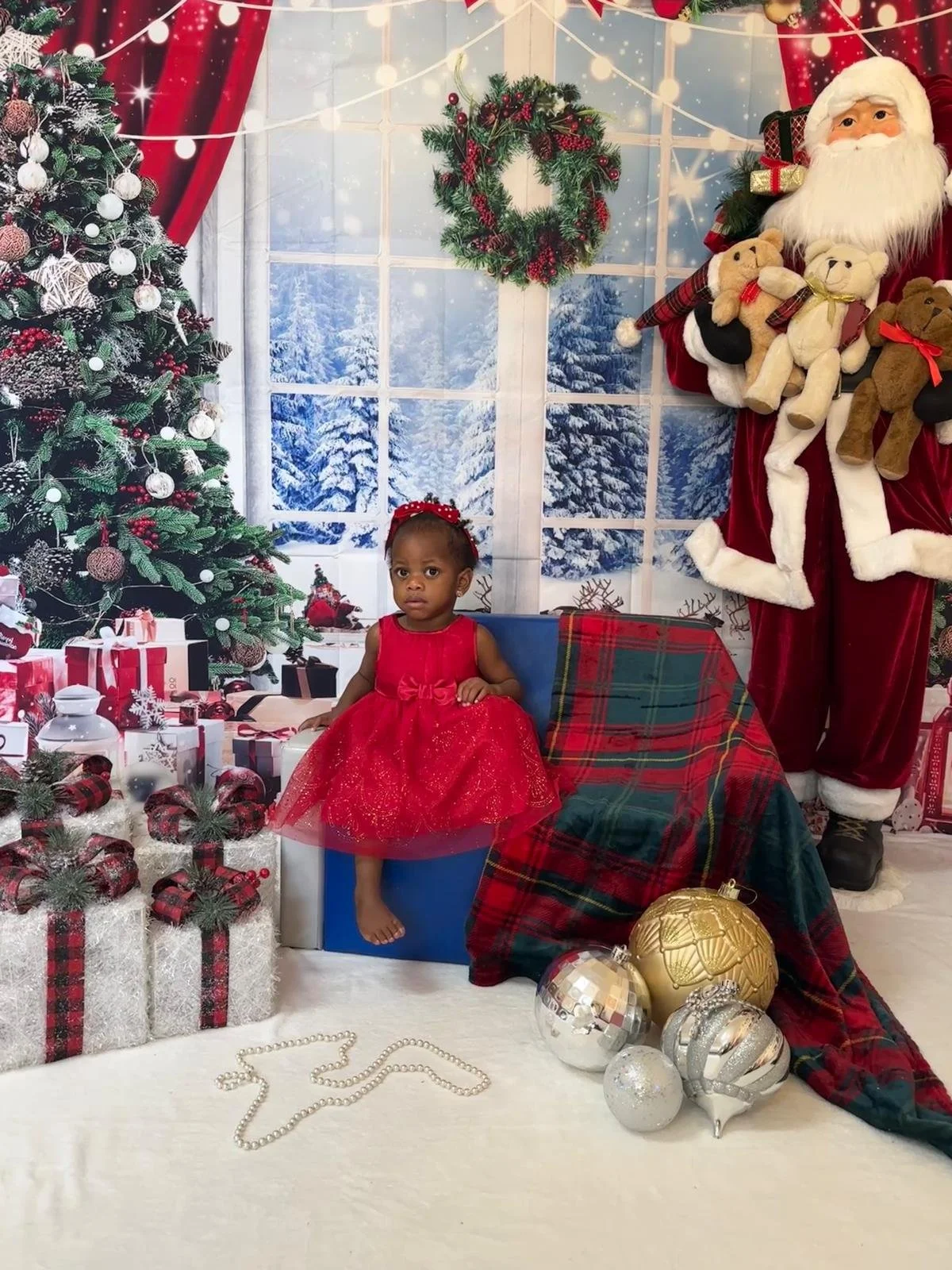 A young girl in a red dress sitting on a box surrounded by Christmas presents, with a decorated Christmas tree and a Santa Claus figure in the background, in a festive holiday setting.
