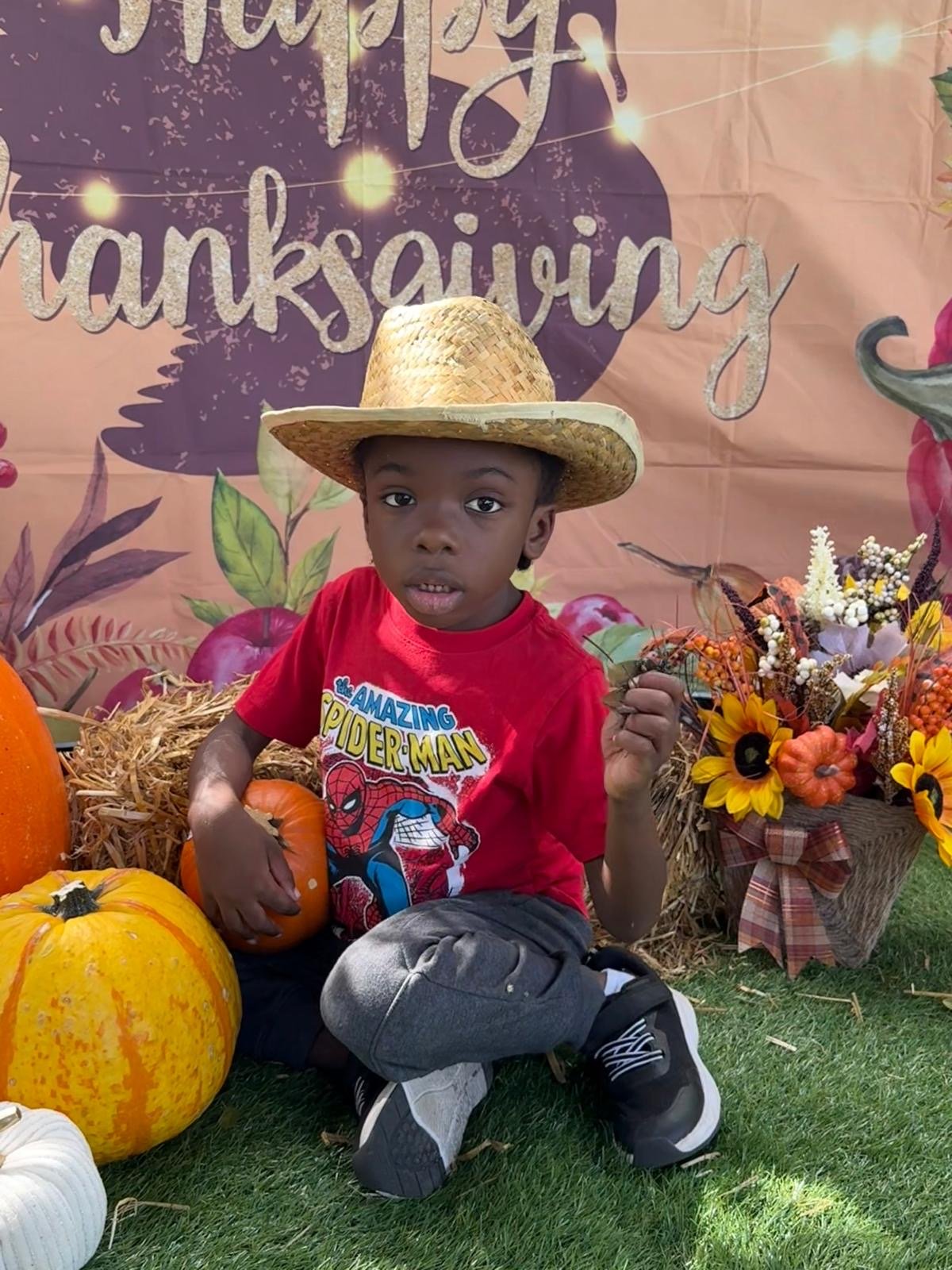 A young boy wearing a straw hat and a red Spider-Man t-shirt sits on grass surrounded by pumpkins and fall flowers, with a Thanksgiving backdrop.
