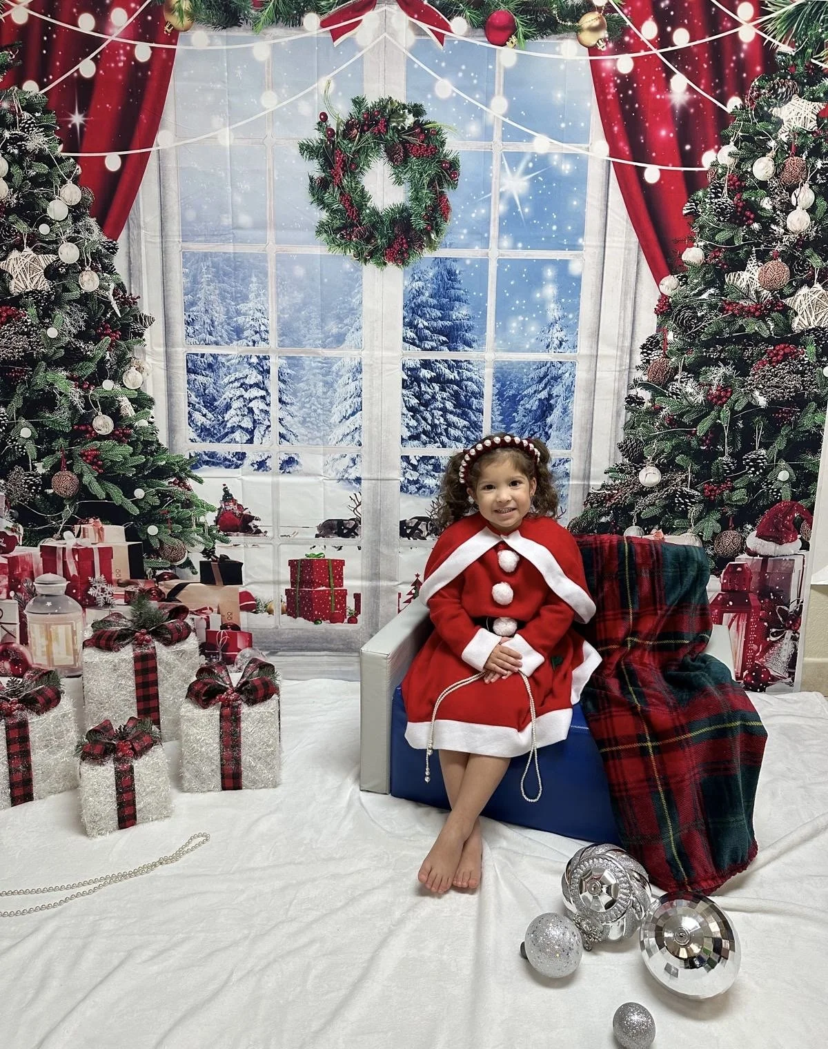 A young girl in a Christmas outfit sitting in front of a festive holiday scene with decorated Christmas trees, wrapped gifts, and snow outside the window.