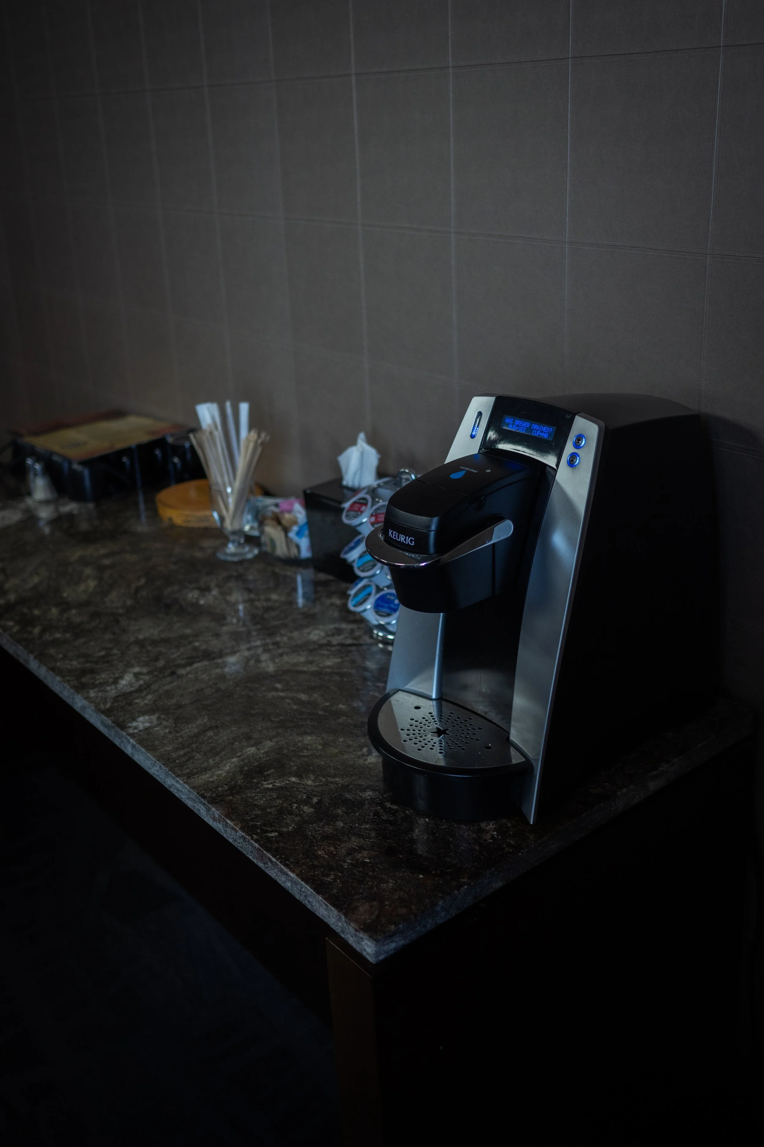 A coffee station with a Keurig machine, stir sticks, napkins, and other supplies on a dark marble countertop.