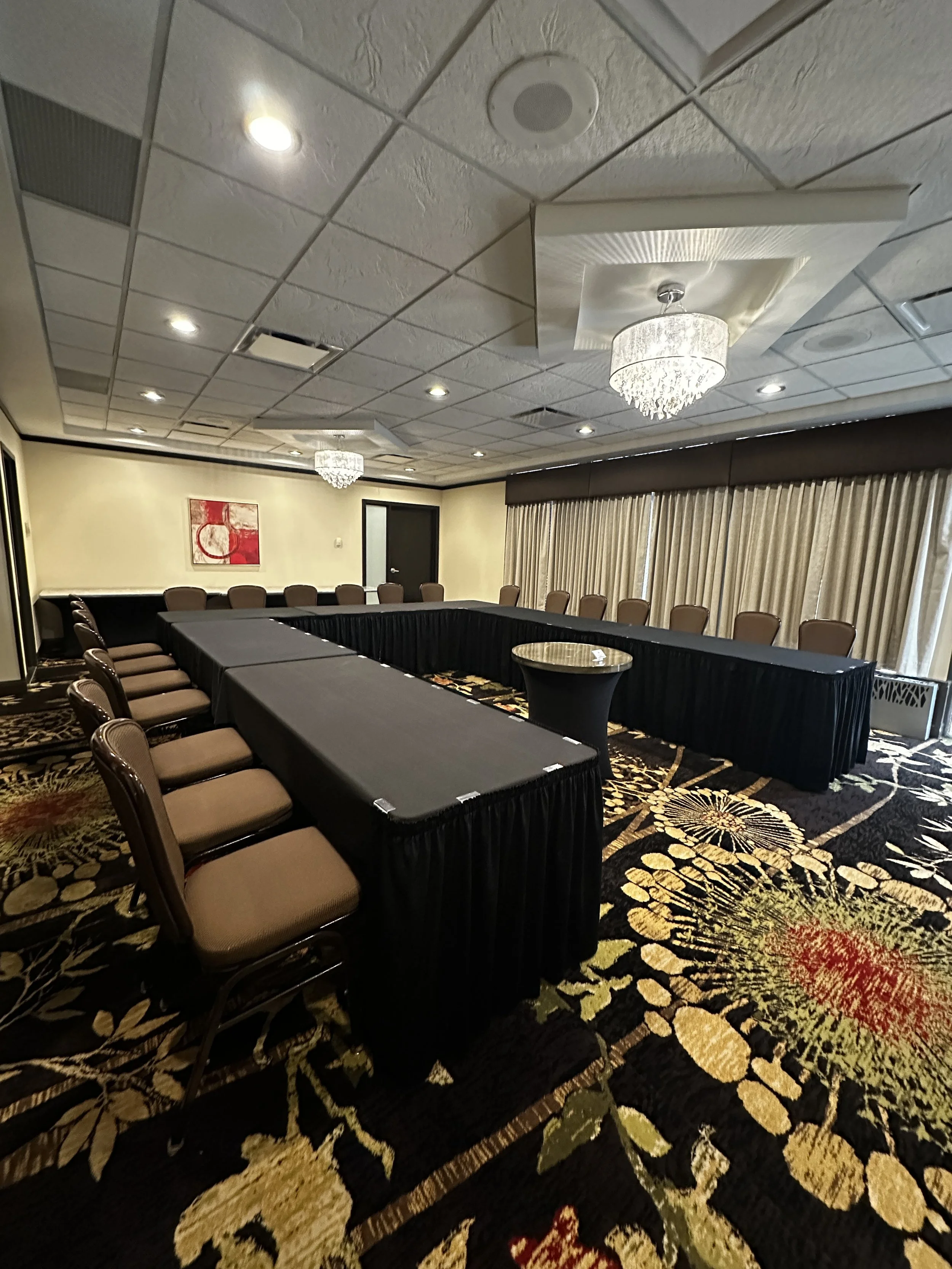 Conference room with U-shaped table covered with black tablecloths, surrounded by brown chairs, chandelier lighting, patterned carpet, and beige curtains.