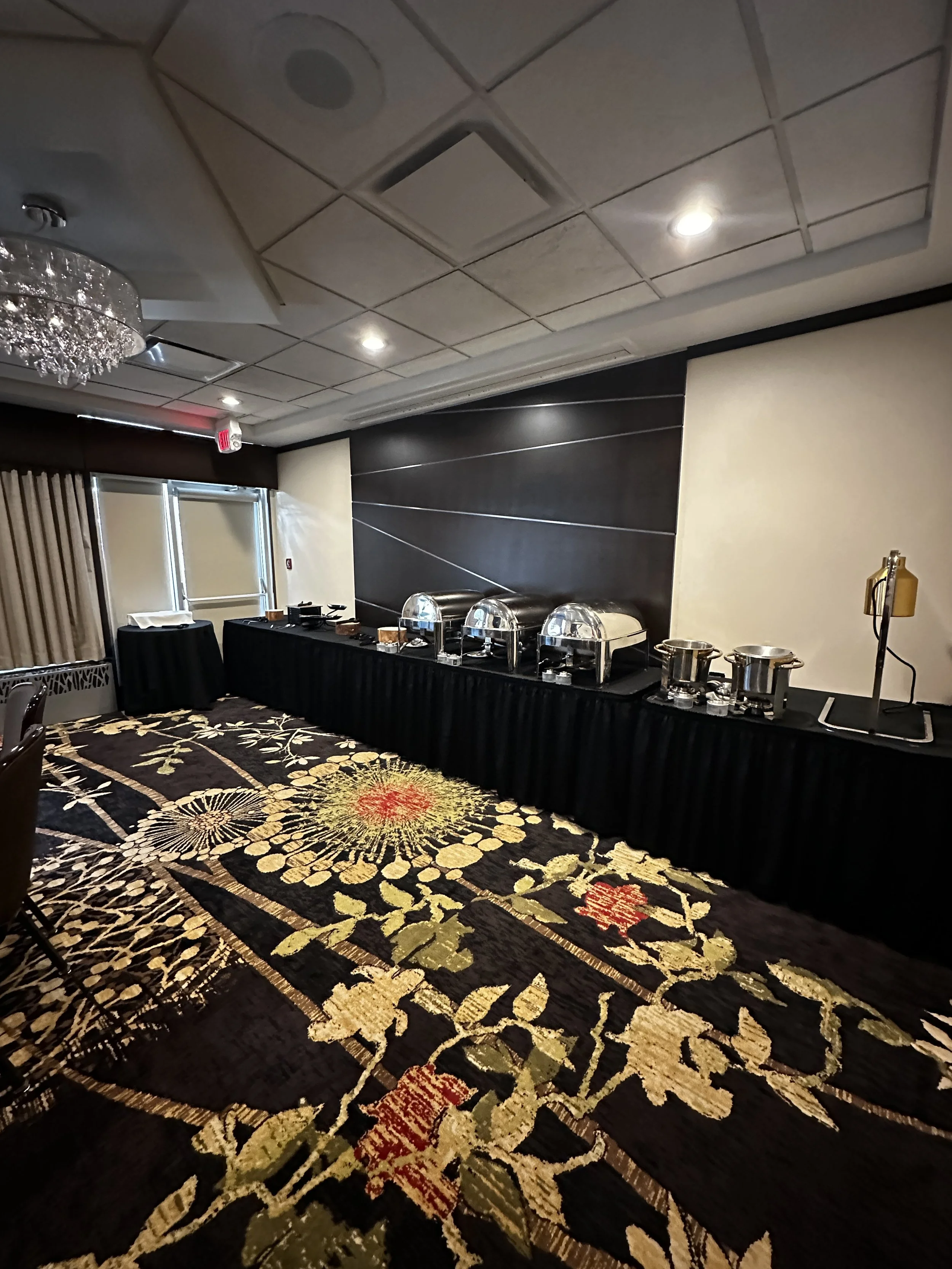 Buffet table with chafing dishes in a hotel conference room, floral patterned carpet, ceiling lights, and a chandelier.