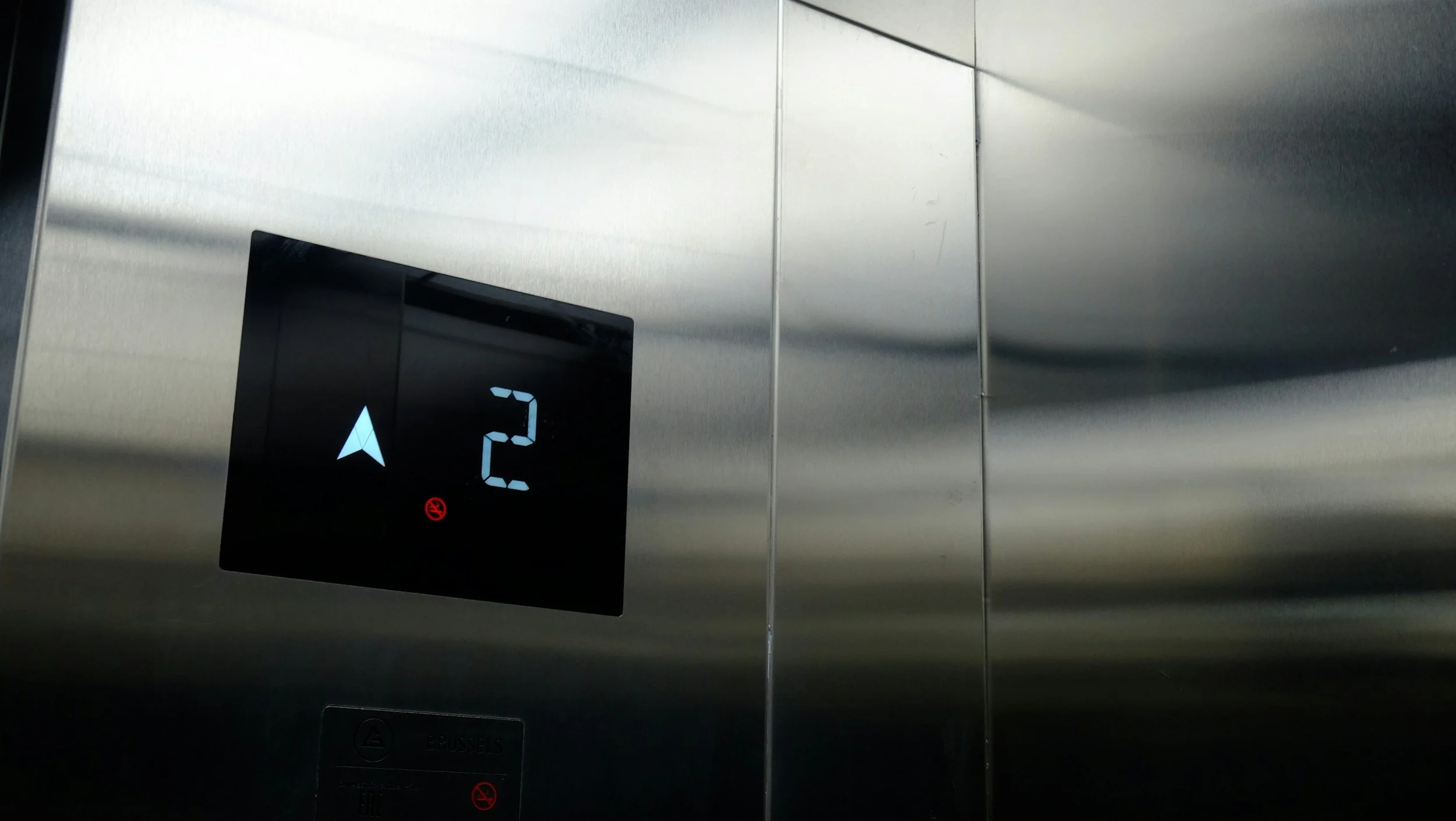 Elevator control panel showing the floor number 2 with a blue arrow pointing up, and a red 'no smoking' sign.