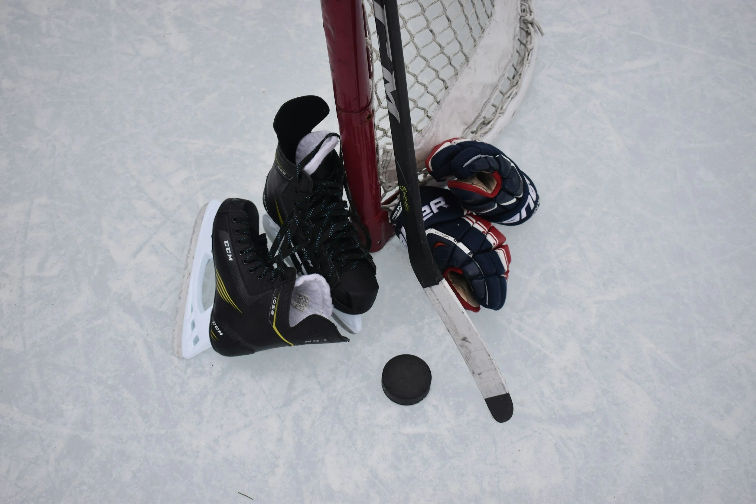 Hockey gloves, skates, a hockey stick, and a puck next to a hockey goal on ice.