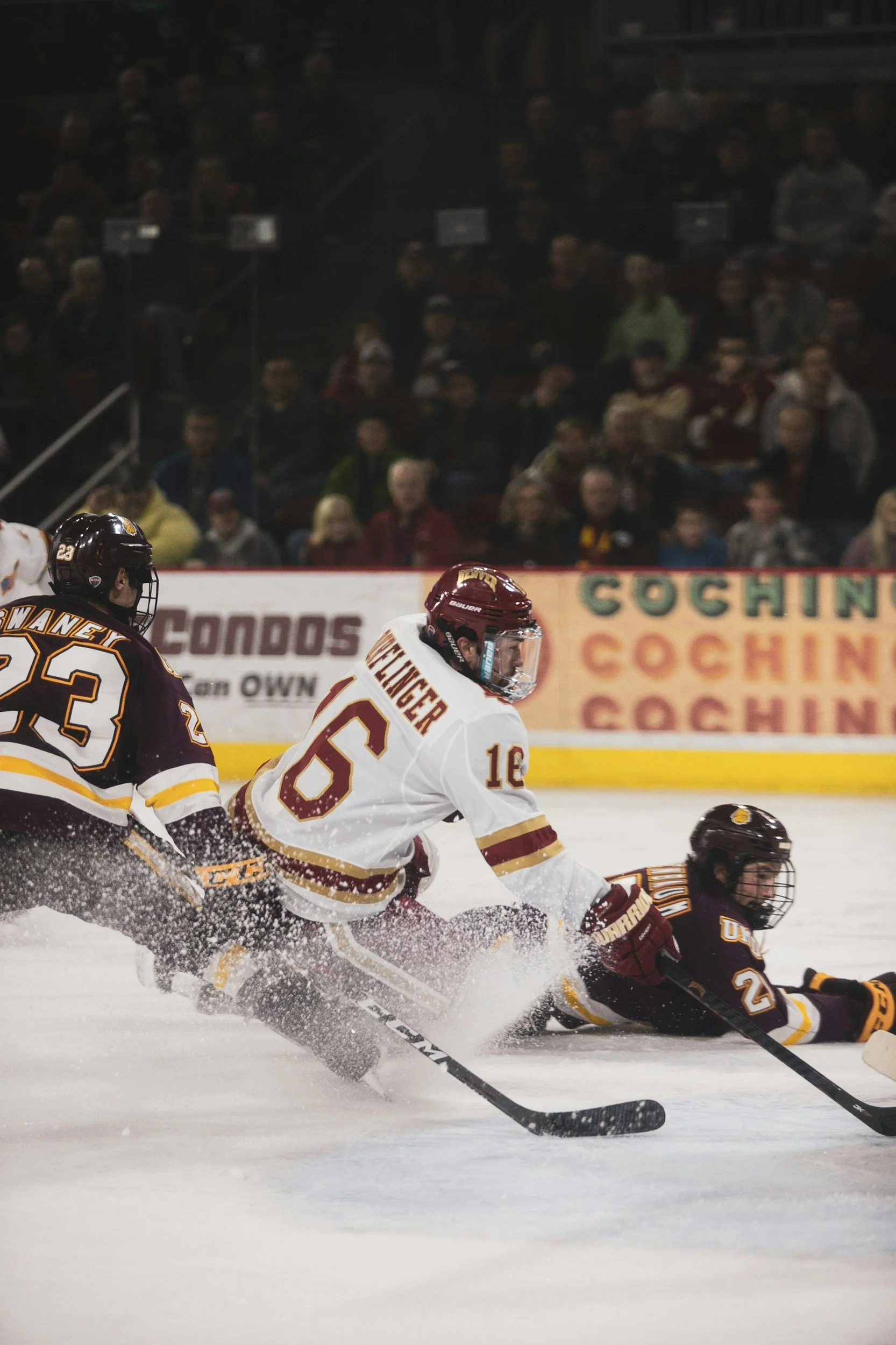 Ice hockey players in mid-play during a game, with some players falling and skimming the ice, and a crowd watching in the background.