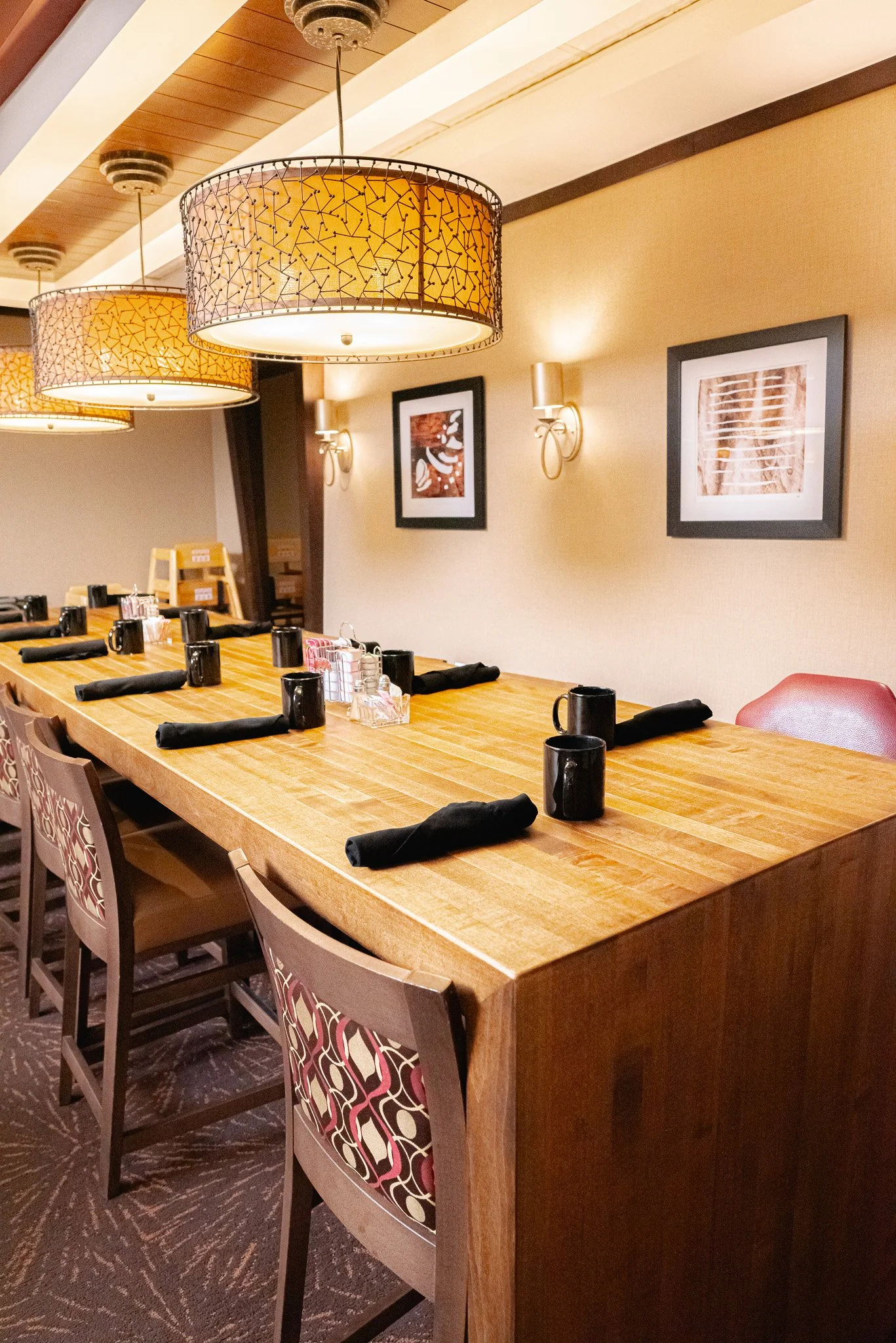 Empty restaurant table with black mugs, black napkins, and condiments, decorated with hanging lamps and framed artwork on the wall.