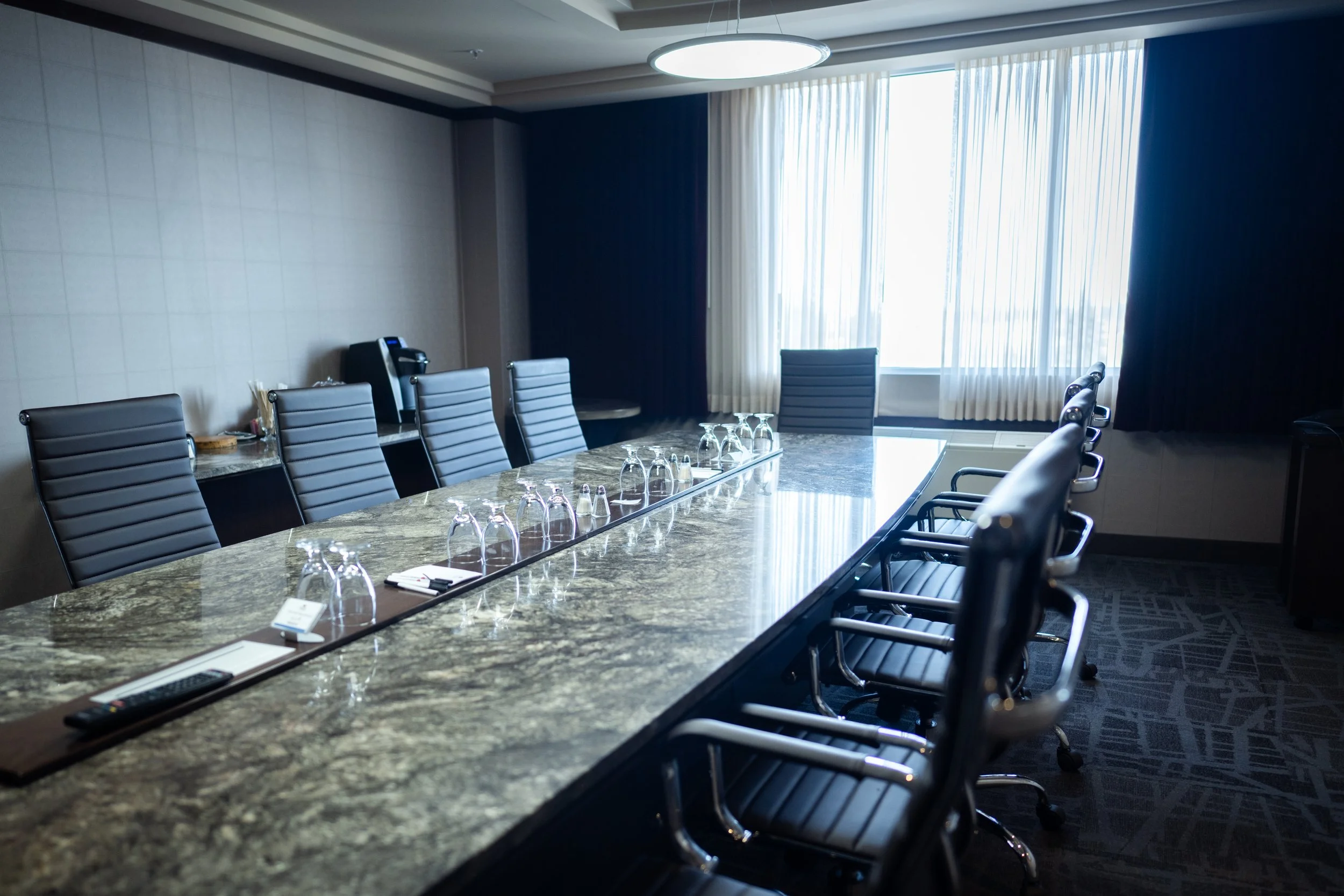 Empty conference room with a large marble table, glassware, and black leather chairs, with large windows and white curtains.