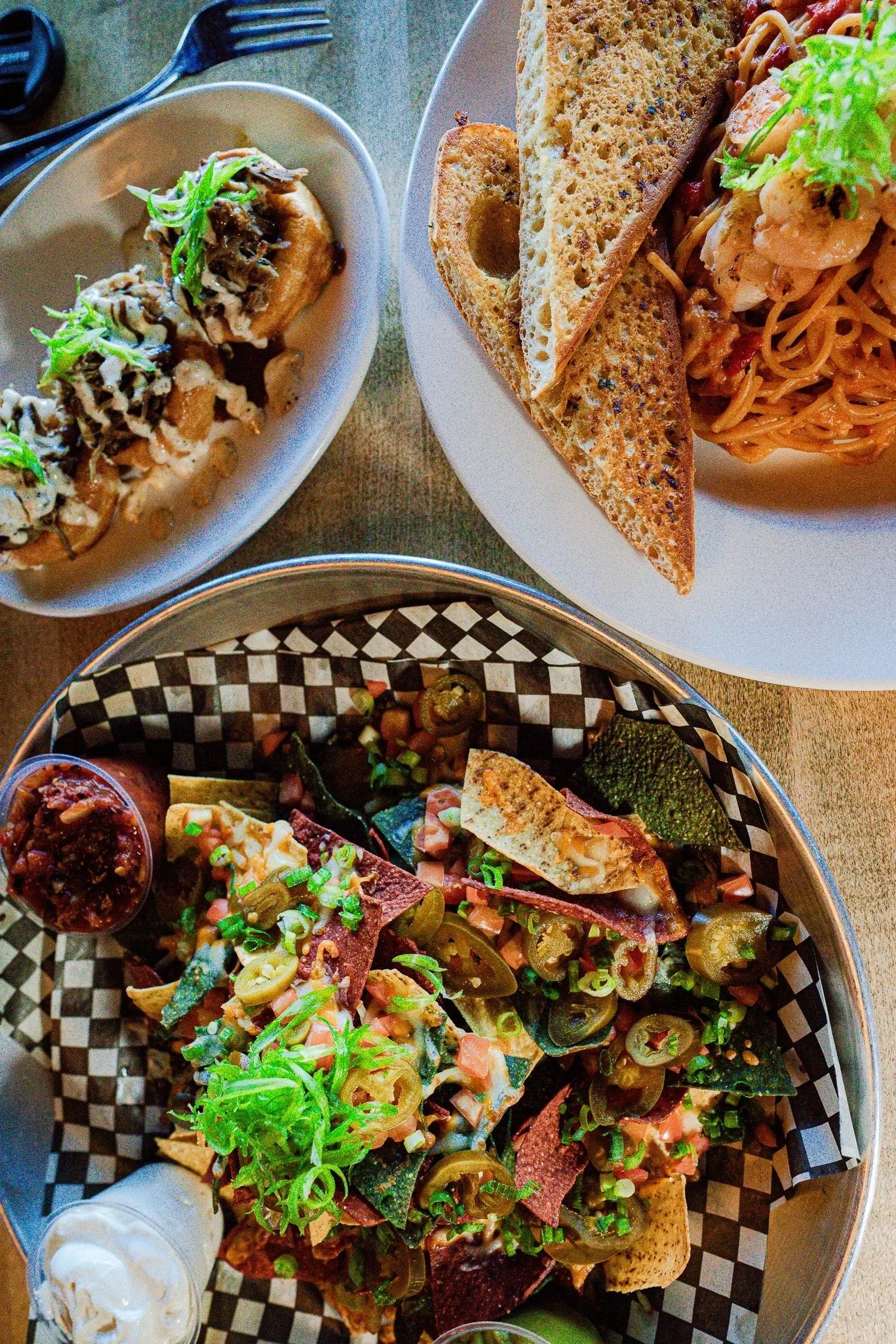A table with various dishes, including nachos topped with jalapenos and chopped green onions, a plate of spaghetti with bread, a small dish of fried food with greens, and a basket of chips and salsa.