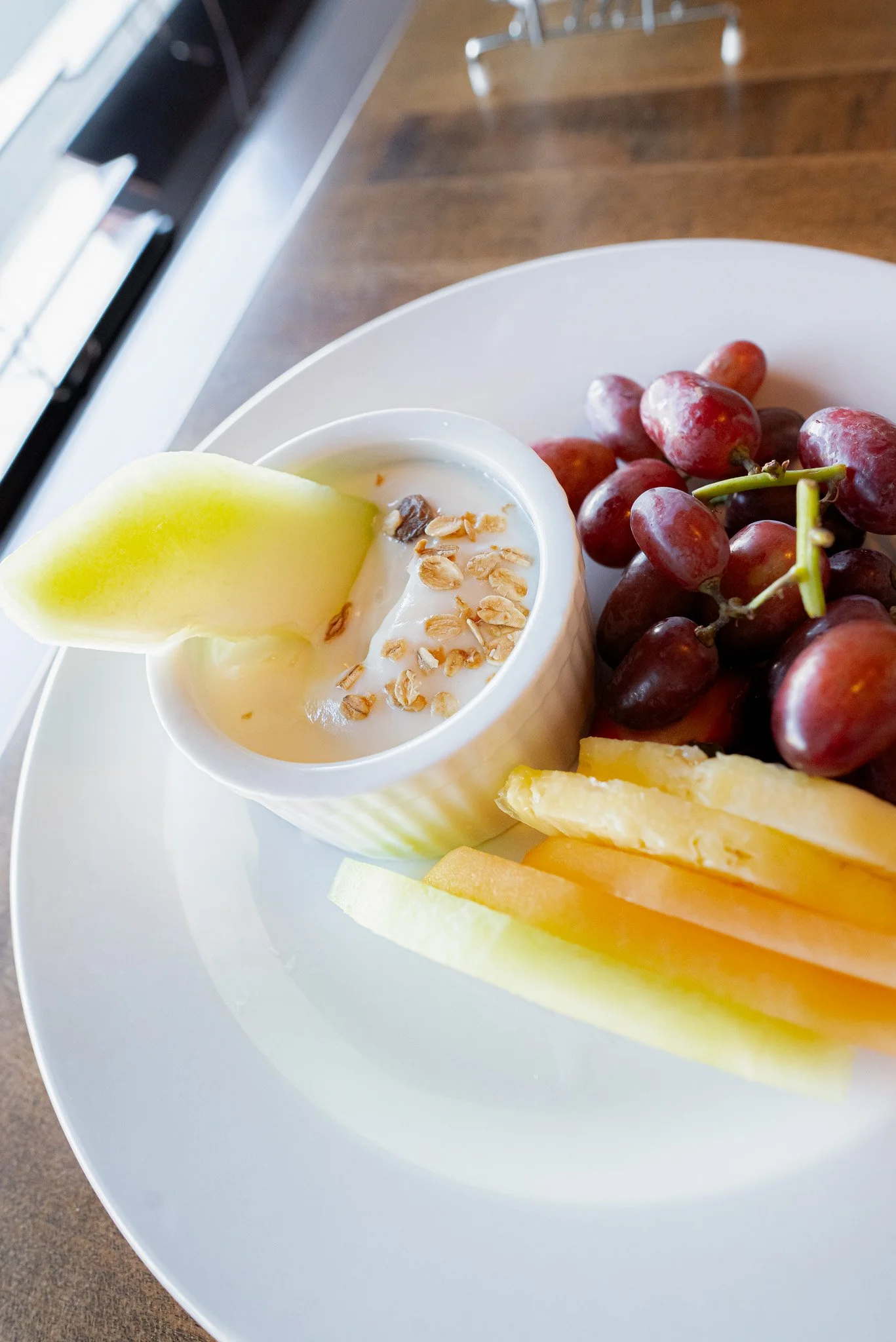 A plate with red grapes, sliced cantaloupe, slices of honeydew melon, and a cup of yogurt topped with oats and a melon wedge.